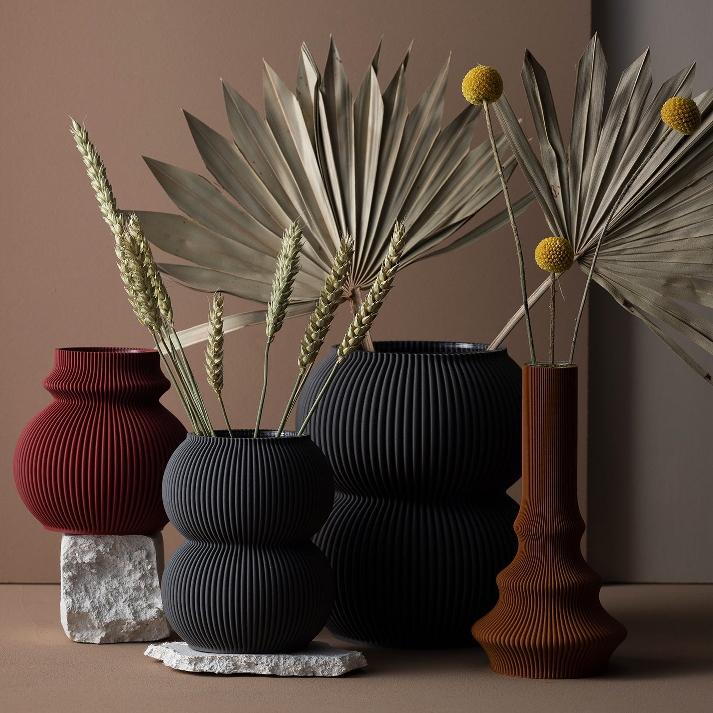 The Zayl 159g Vase by Sheyn is crafted from recyclable materials and made through 3D printing. Available in various shapes and earthy tones, including red, black, and brown, it displays dried plants like palm leaves and billy balls against a neutral background with stones underneath.