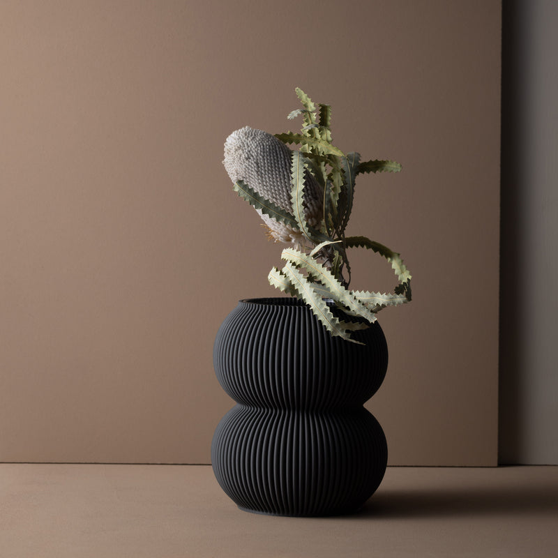 The Zayl 130g Vase by Sheyn, a textured, 3D-printed double-sphere vessel, houses a unique cactus with a round, fuzzy top and long, wavy leaves. Set against a muted brown backdrop, this eco-friendly arrangement is minimalist and monochromatic.