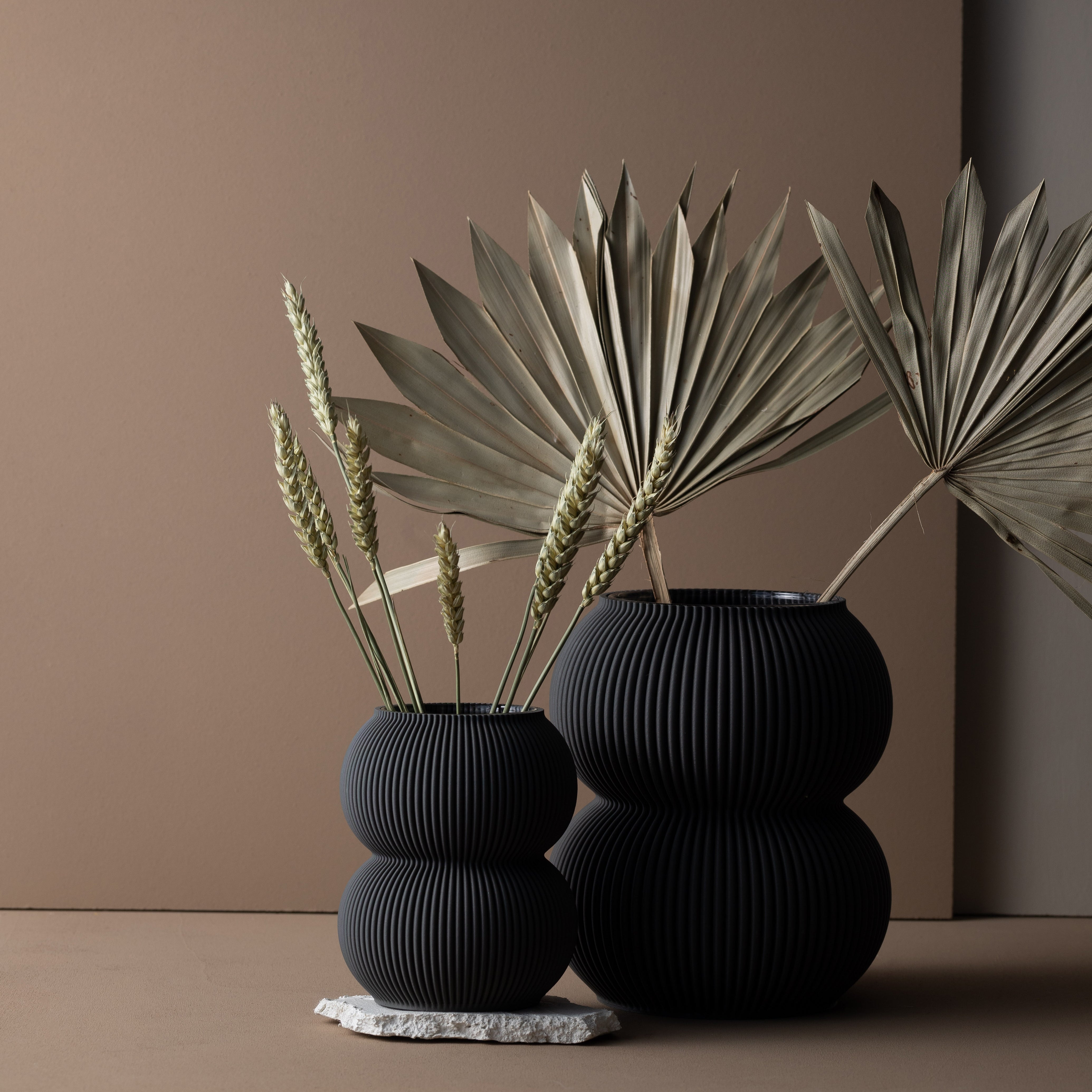 Two black vases from Sheyns Zayl 159g collection, made from recyclable materials, rest on a neutral background. The small vase holds wheat stalks and is slightly elevated by a small stone, while the larger contains dried palm leaves, creating a minimalist natural arrangement.