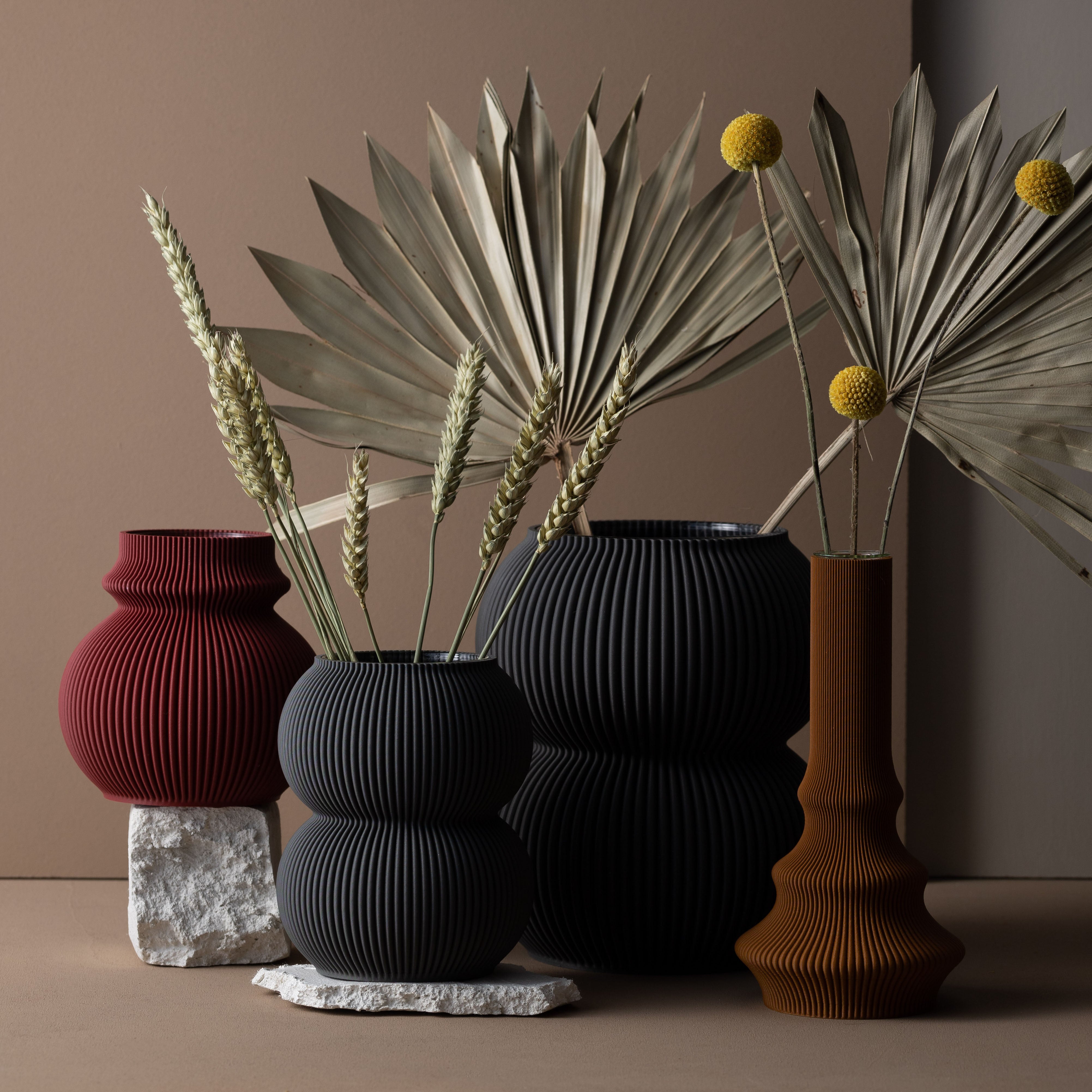 Sheyns Zayl 130g vase collection, featuring eco-friendly ribbed designs in red, black, and brown, showcases dried wheat stalks and desert flora. Artistically arranged with white stone accents on a neutral background, they resemble elegant architectural pillars that echo natures beauty.