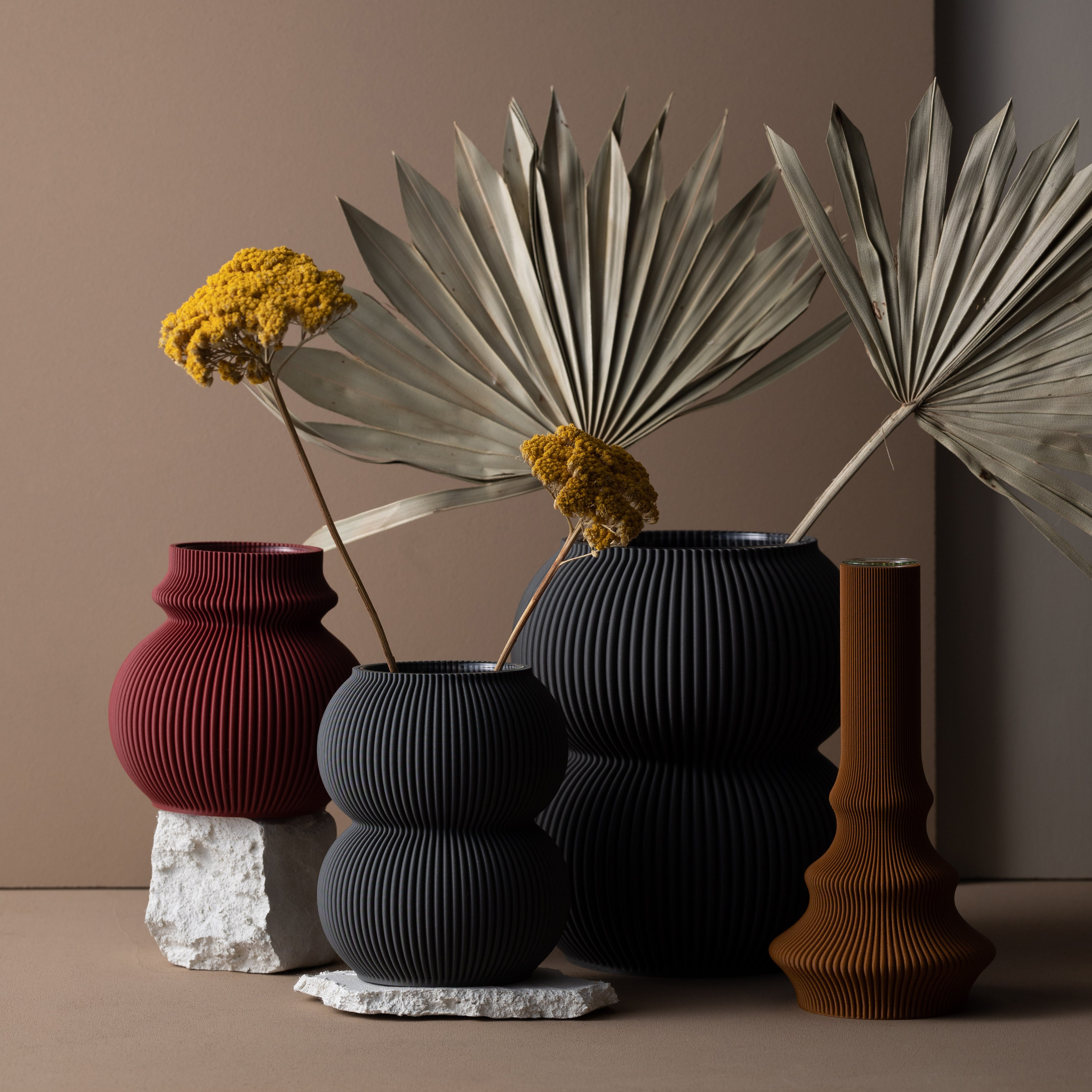 The Zayl 114g Vase by Sheyn is a ribbed ceramic piece in earthy tones of deep red, black, and brown. Crafted with an eco-friendly approach, it holds dried palm leaves and yellow flowers against a minimalist beige backdrop with stone accents.
