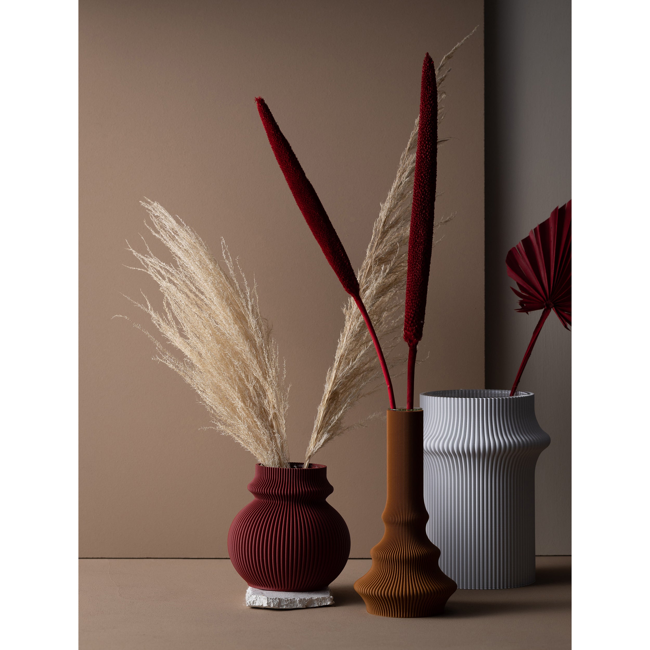 A minimalist display features the Zayl 114g Vases by Sheyn, in brown and gray tones, made with eco-friendly materials. They hold pampas grass and dark red dried flowers against a neutral beige backdrop for an elegant, earthy contrast.