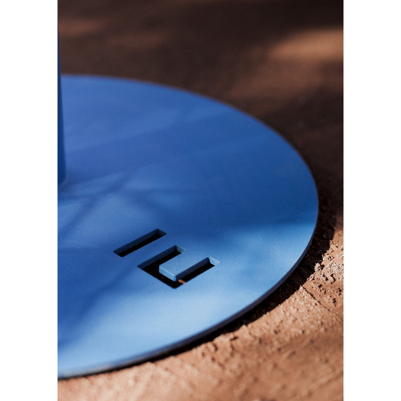 Close-up of the Spulka Table by Egoe, featuring a blue circular metal base with two rectangular cutouts casting a shadow on a textured brown surface, highlighting its durable all-metal body.
