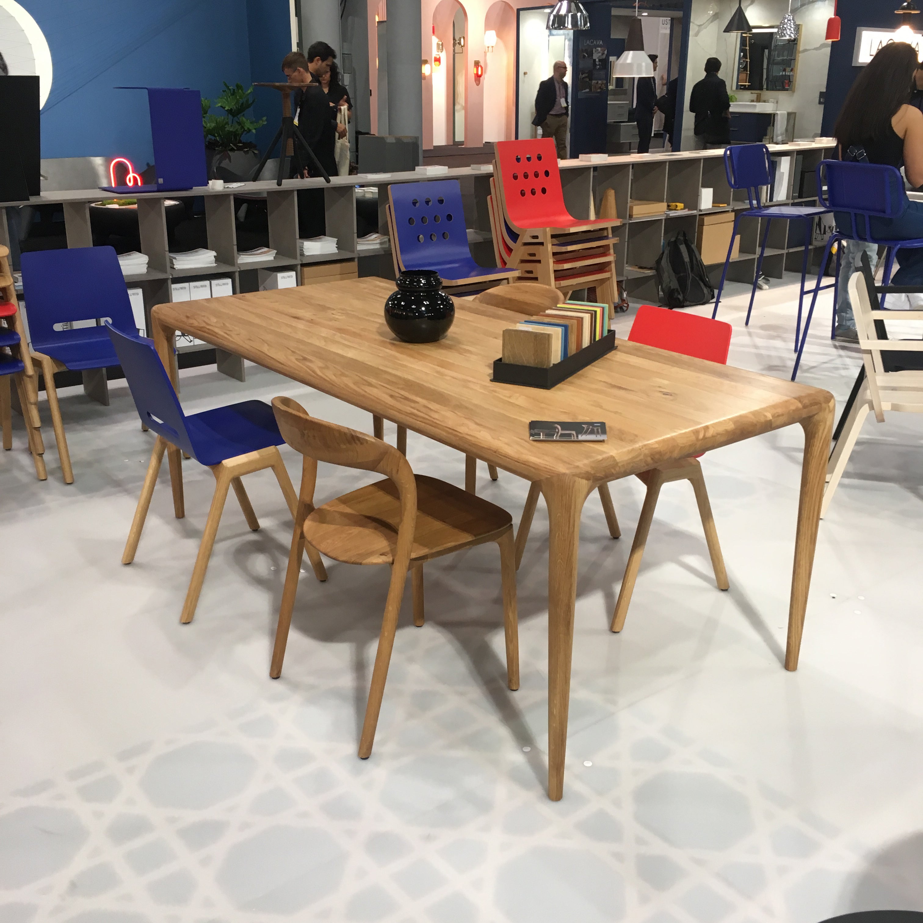 At a trade show, the Forum 2S - Ultramarine Sale by Hussl showcases a modern wooden dining table with curved edges. Its surrounded by stackable timber chairs in red and blue. The tabletop displays a black vase and colorful material samples, while people are visible in the background.