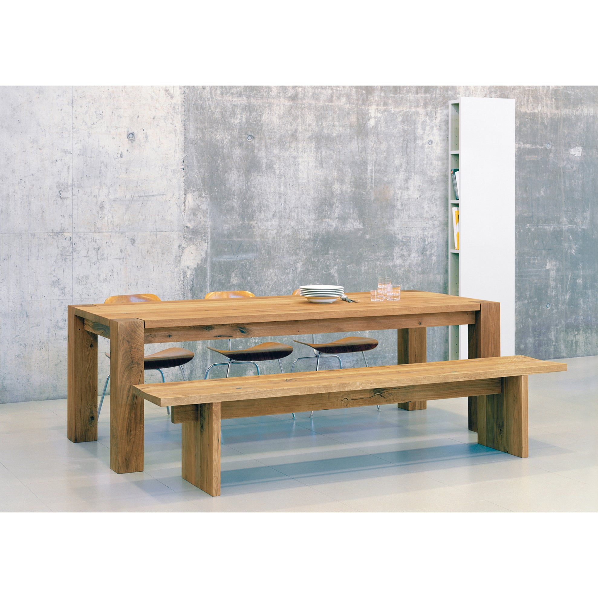 The dining area includes a large wooden table with E15s designer flair, complemented by the BE02 TARO bench and three wooden chairs. A textured gray wall serves as the backdrop, while a sleek Philipp Mainzer shelving unit enhances the corners elegance.