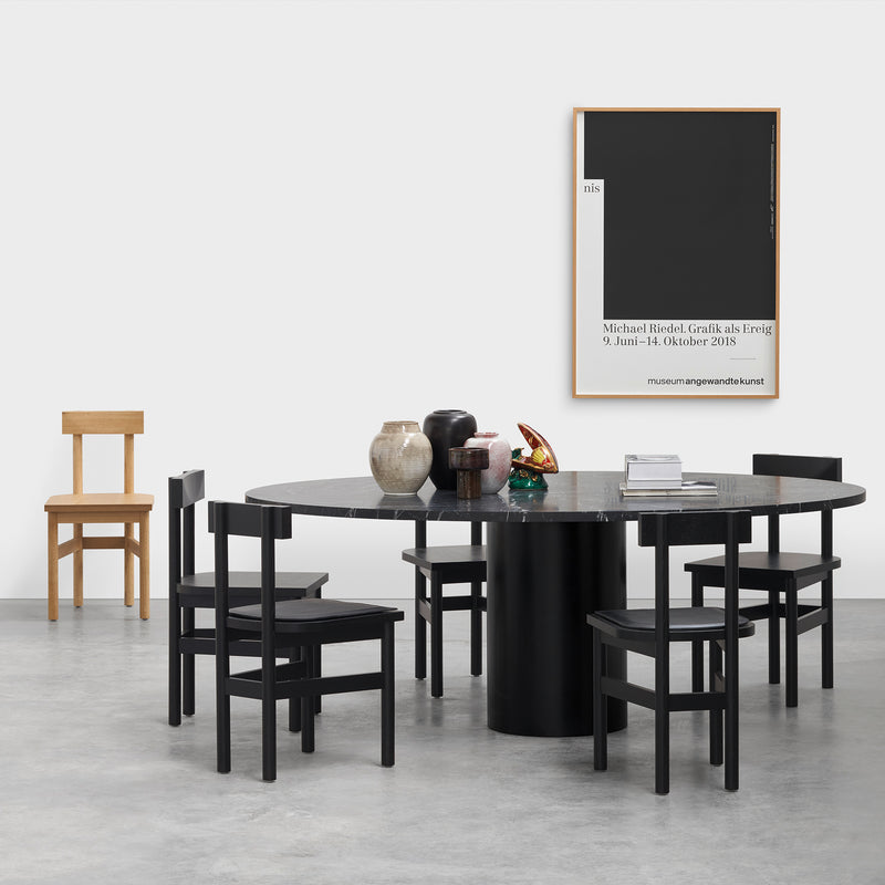 A modern dining area showcases a round black table adorned with vases and candles, flanked by four black chairs. An E15 SX01 GAMAR Chair adds a rustic touch in the background. A minimalist black and white poster decorates the white wall, while the light gray floor enhances the setting.