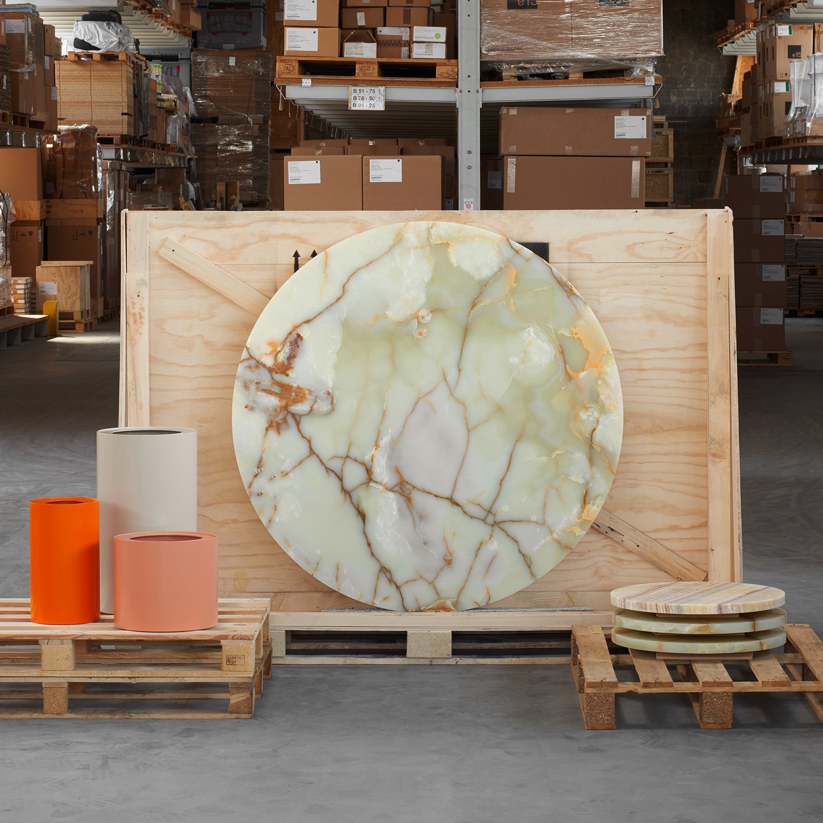 A large, TA20 HIROKI ONYX slab by E15 displays a white and brown marble-like pattern as it leans on a wooden crate. Nearby, cylindrical and round objects in orange and beige are arranged on pallets in a warehouse.