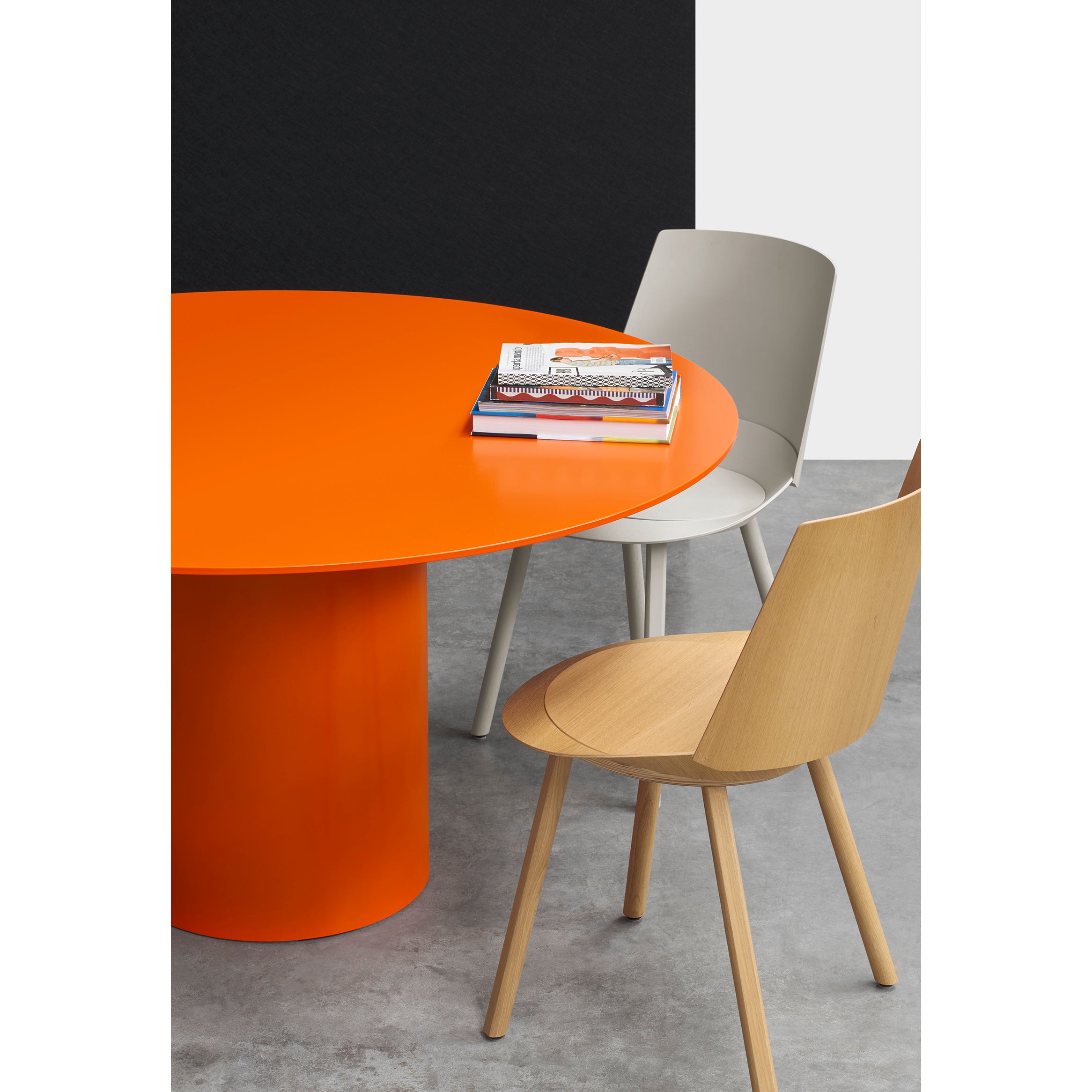 In a modern setting, the E15 TA20 HIROKI METAL table with an orange circular design exudes sculptural quality. Its paired with a light gray chair and wood-toned companion on a gray concrete floor, accented by magazines. A black and white wall adds contrast to this artistic scene.