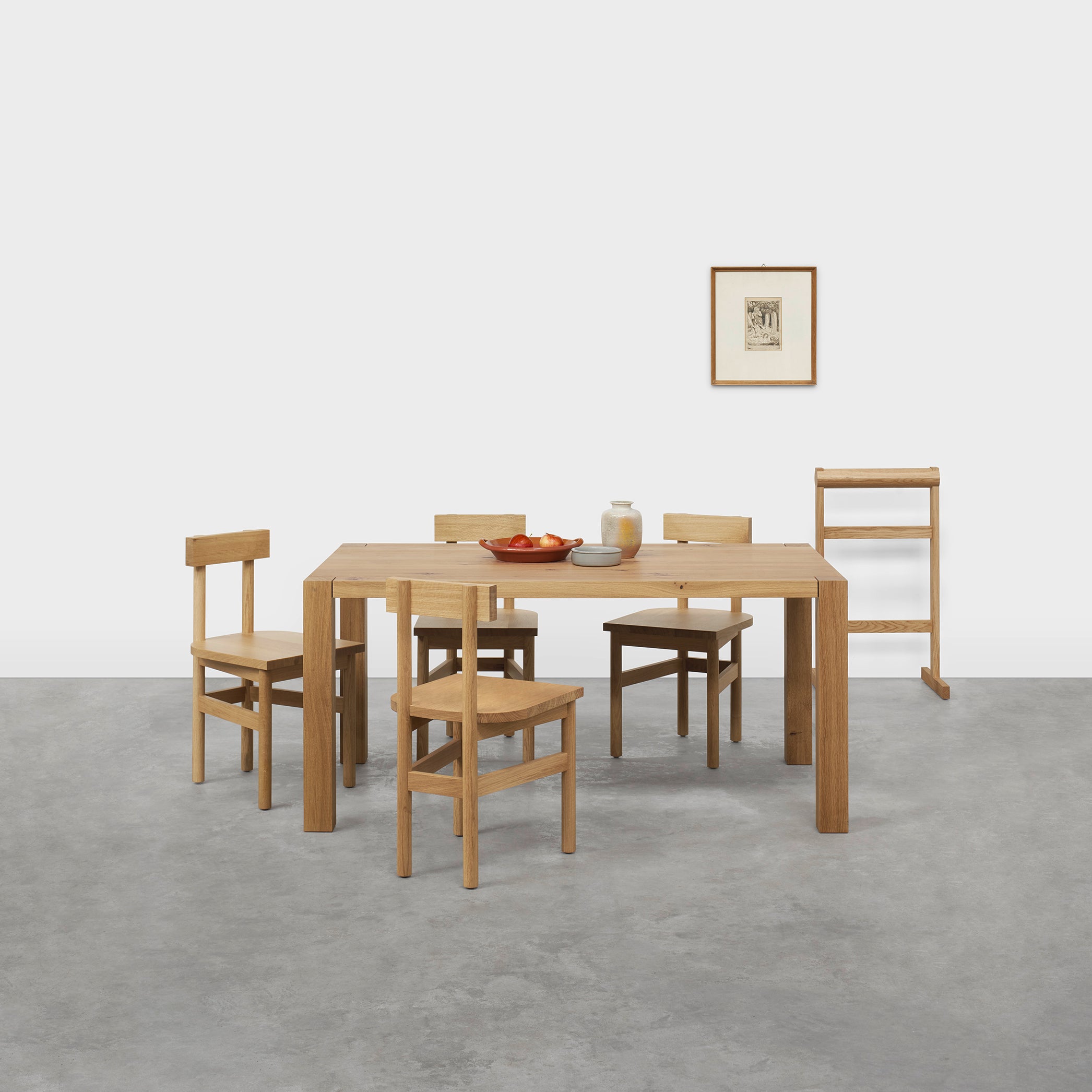 A minimalist dining area features a wooden table with four E15 SX01 GAMAR chairs. A bowl of fruit and a jar rest on the table, while a framed picture enhances the white wall. Nearby is a narrow shelf or towel rack, and the concrete floor completes the simple yet charming look.