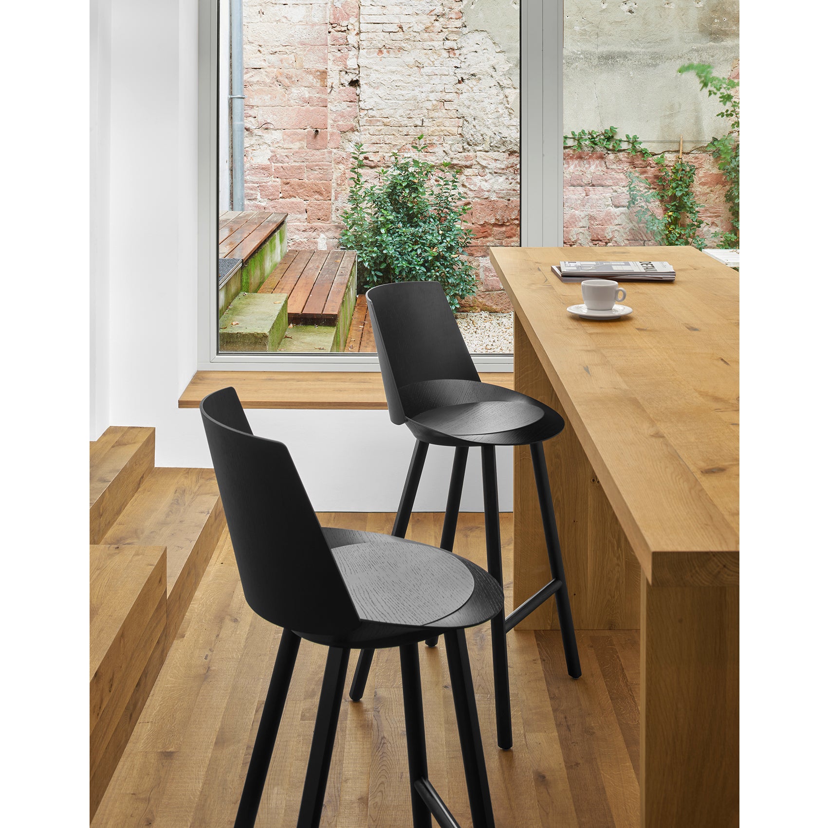 Two E15 ST10 JEAN With Backrest black bar stools adorn a light wooden counter in a modern setting, elevating the elegant dining experience. A cup and saucer are on the counter, with a window view of a brick wall, wooden steps, and lush greenery behind it.