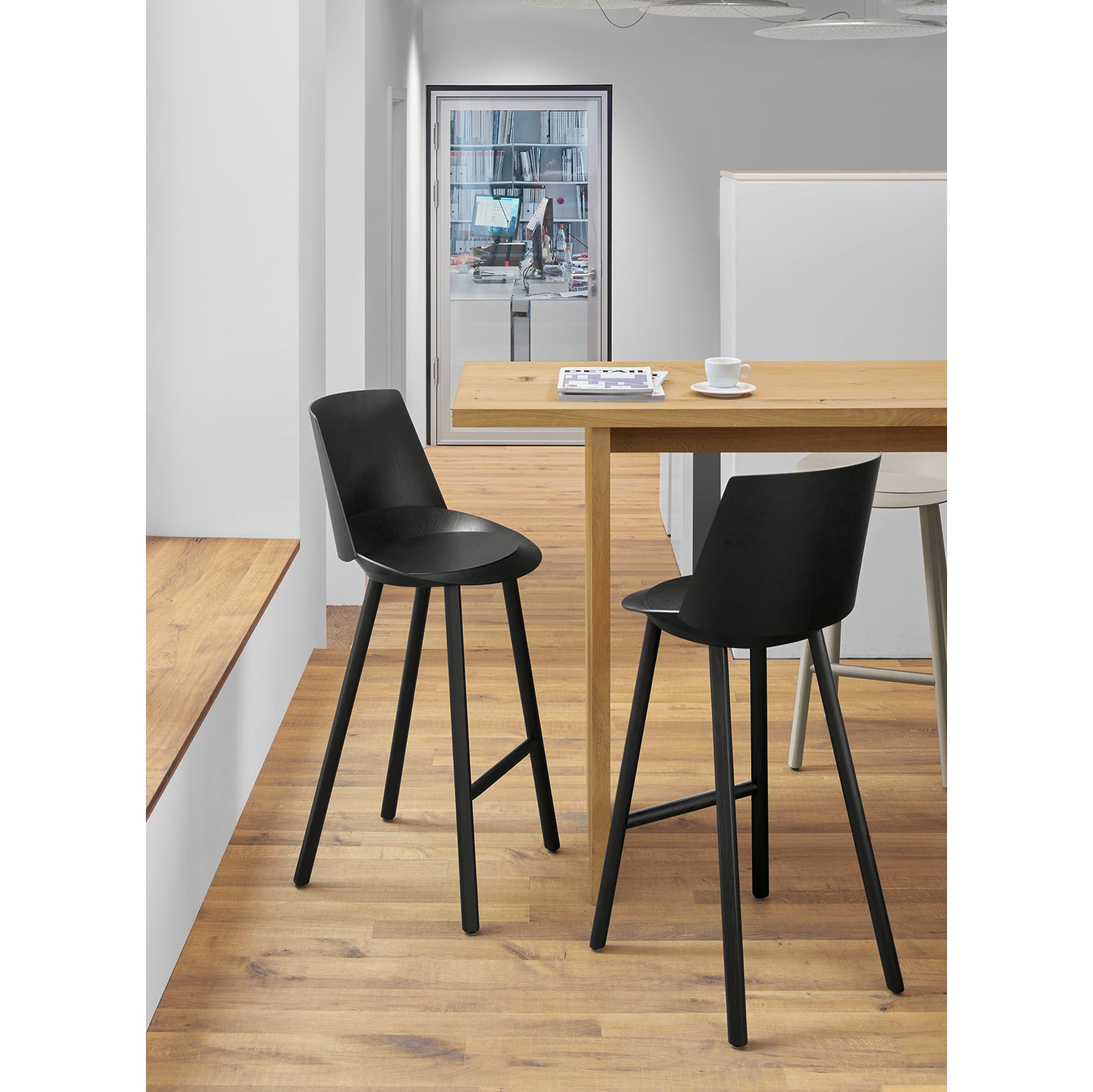In a modern office, E15s ST10 JEAN With Backrest delivers ergonomic comfort around a wooden high table on a light wood floor, paired with two sleek black stools. A white coffee cup and magazine add an inviting touch, while the glass-walled room enhances the bar-dining ambiance.