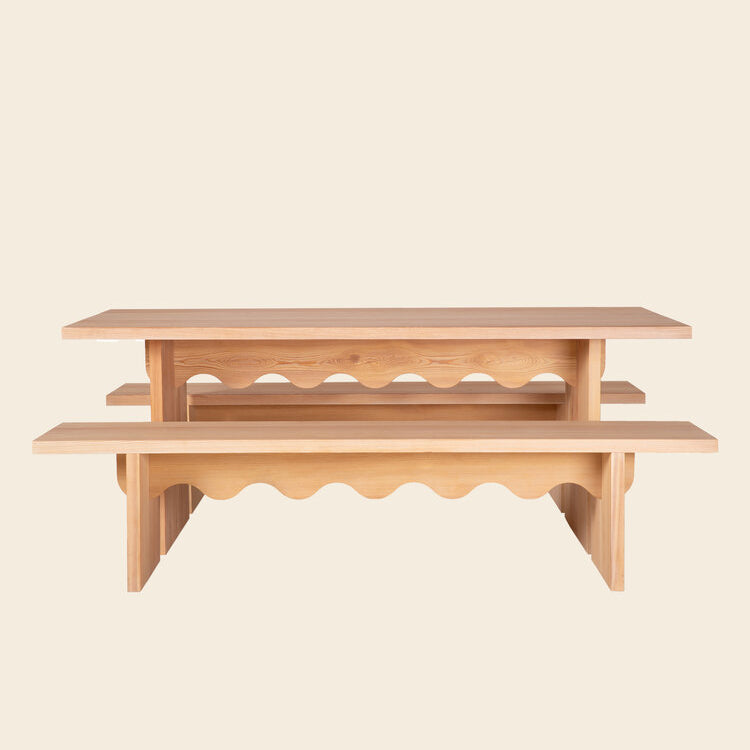 The Tami Dining Collection by Julia Jessen, crafted for Schneid, features a wooden set made from naturally oiled larch wood. It includes a Tami Table and two matching benches with wavy cut-out side patterns, elegantly displayed against a neutral backdrop.