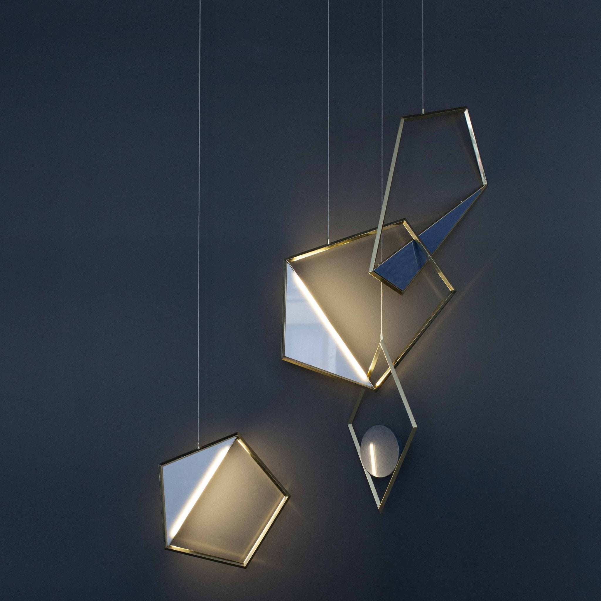 The Tangle Light by Coordination Berlin features sculptural pendant lights with gold frames and integrated LEDs, set against a dark background. These geometric designs, resembling modern jewelry with pentagons and diamonds, create an artistic ambiance.