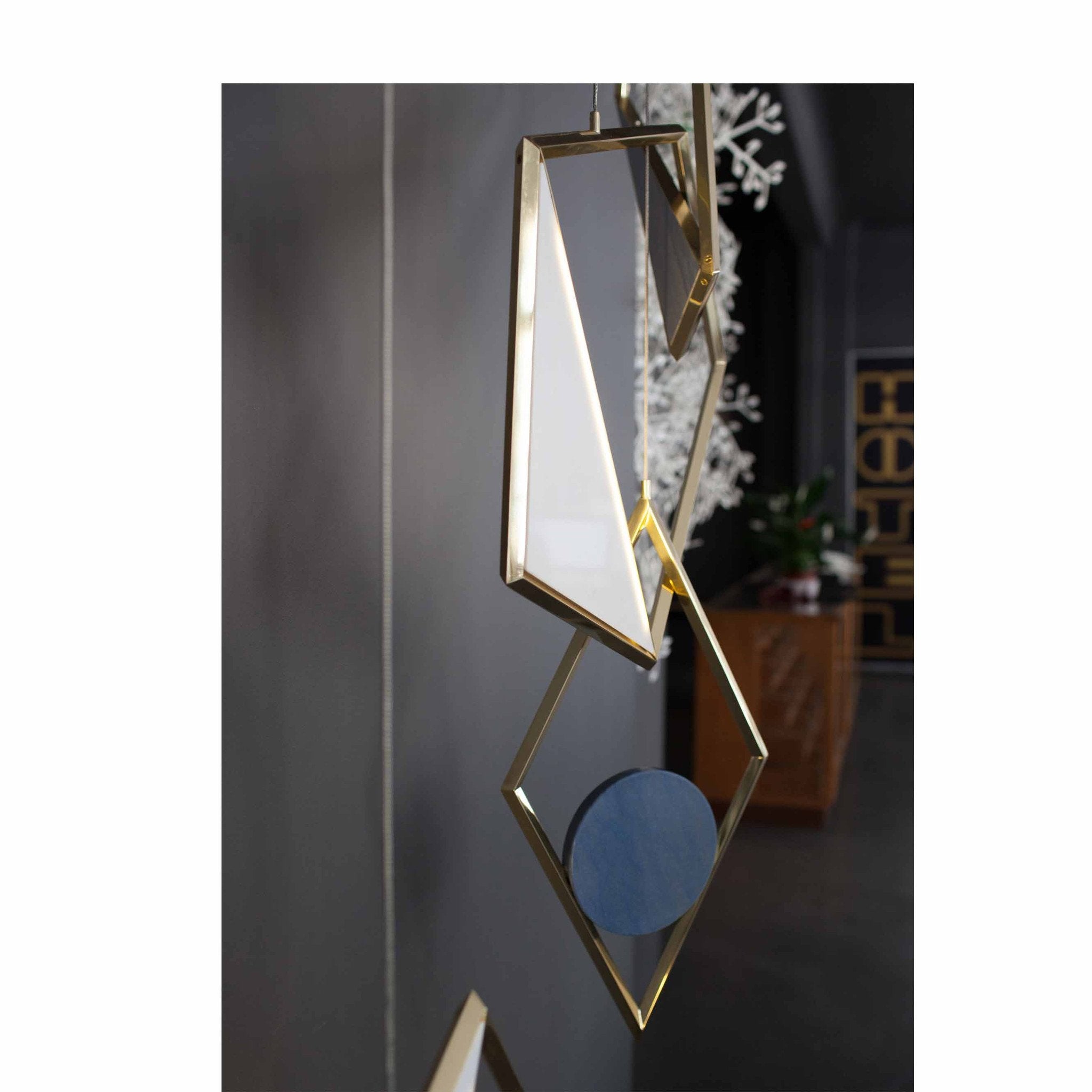Coordination Berlins Tangle Light and geometric gold-framed sculptures, featuring diamond shapes with reflective blue surfaces, hang vertically against a dark wall. The decor suggests a modern, artistic interior inspired by contemporary jewelry design.
