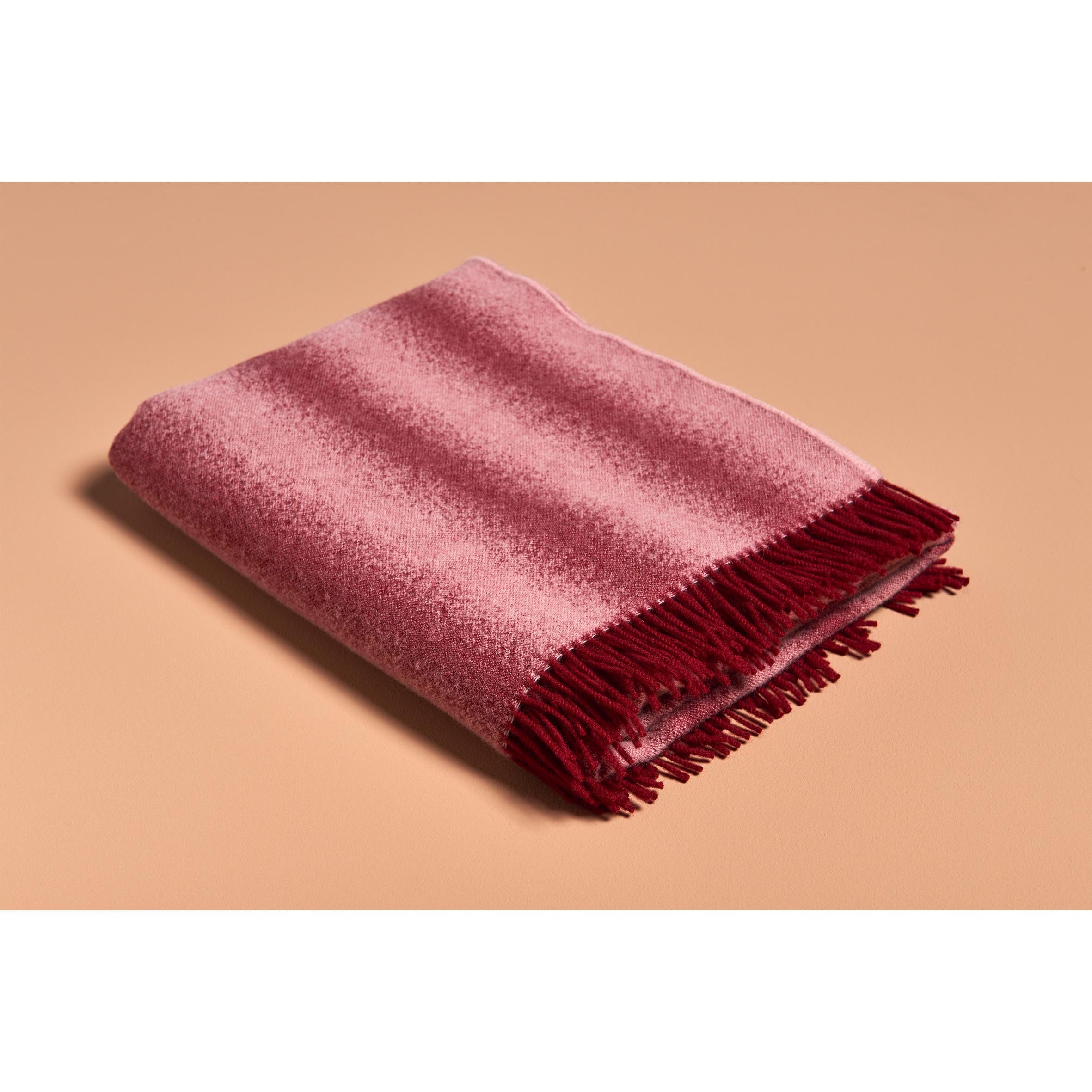 The Schneid Tide Blanket, made from 100% wool and featuring dark red fringes, is elegantly folded on a beige surface. Its pink fabric enhances the ambiance with a soft, textured appearance.