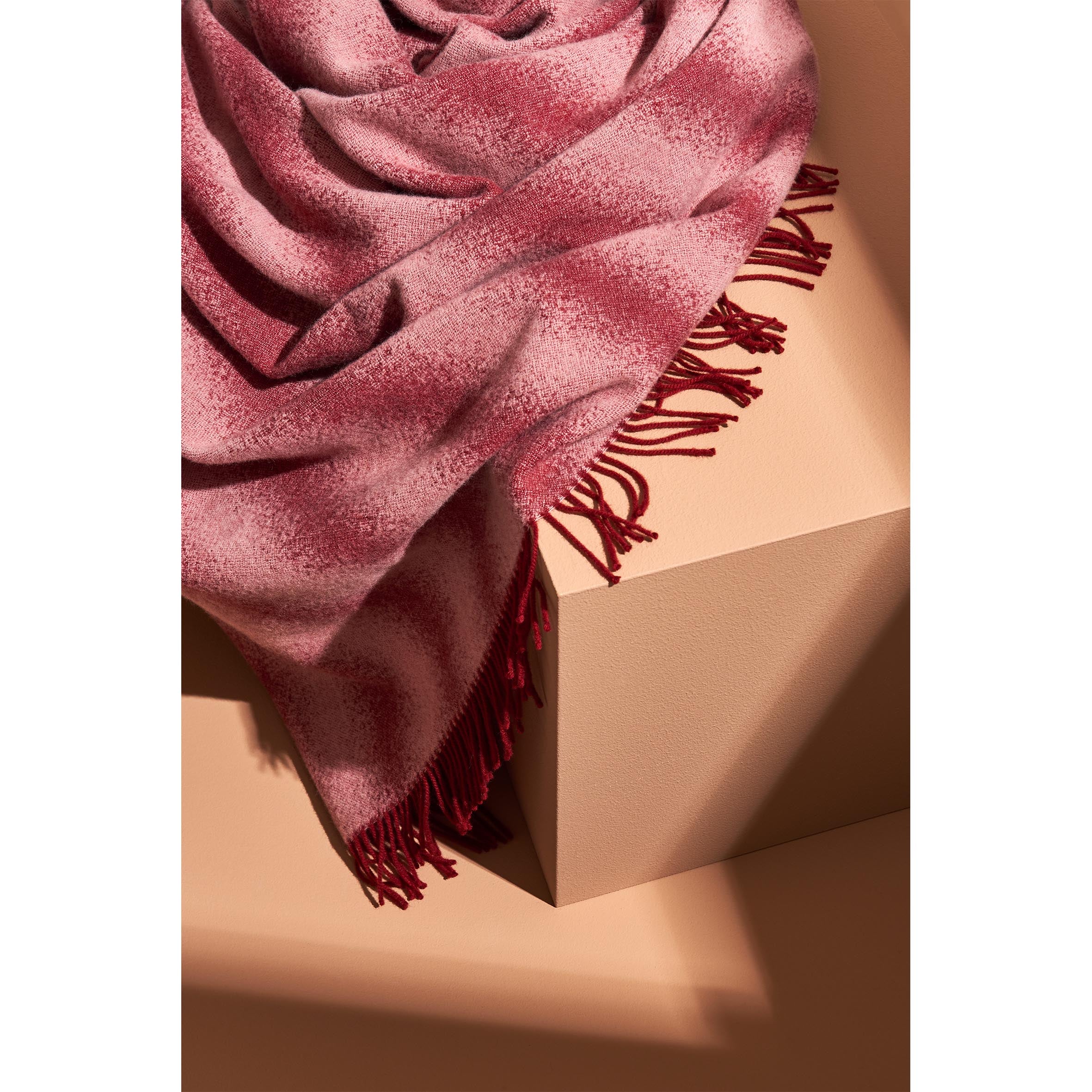 The red and white speckled Tide Blanket from Schneid, made from 100% wool and featuring fringe, drapes over a light brown geometric block. Its texture and color add warmth against the neutral background.