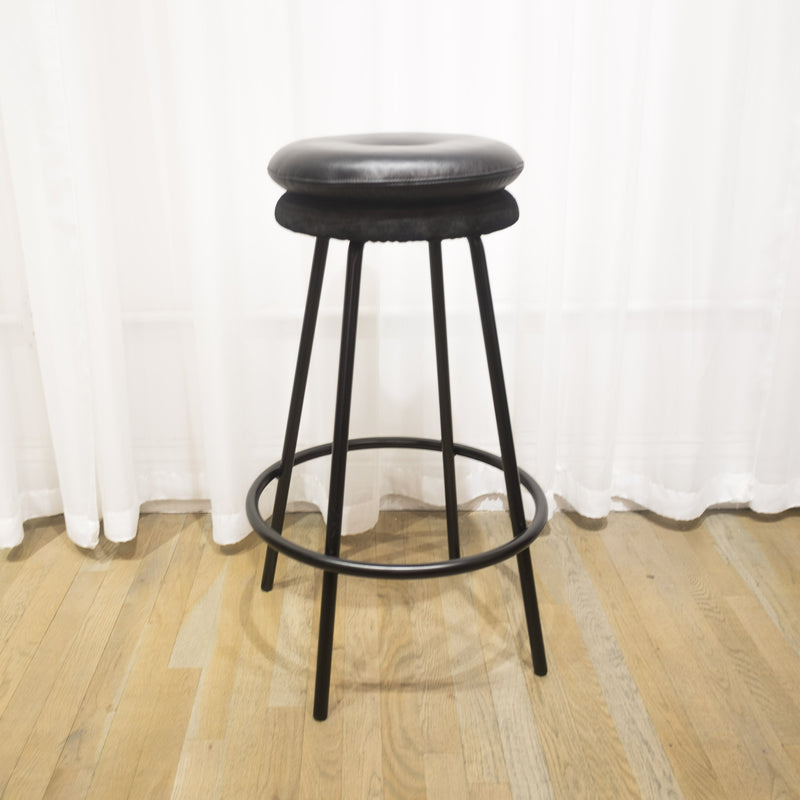 The Big Tom Barstool by Richard Lampert features a sleek black cushioned round seat with a tubular steel frame and straight legs, set on a wooden floor against a white curtain backdrop. Ideal for kitchen counters, its circular footrest enhances both style and comfort.