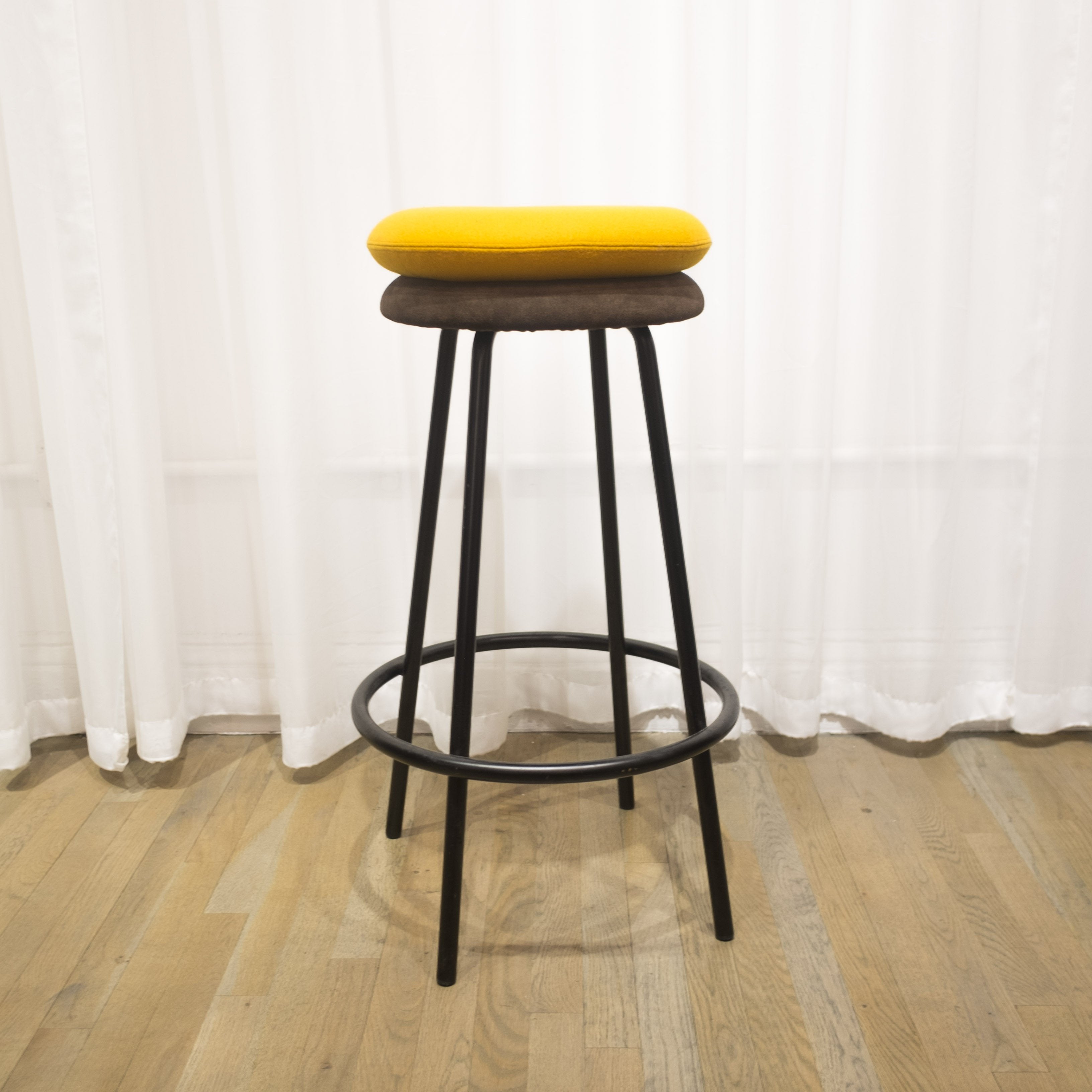 The Big Tom Barstool by Richard Lampert showcases a tubular steel frame, enhanced by two cushions—one in vibrant yellow and the other in rich brown leather. It complements wooden floors beautifully and stands out against pristine white curtains.