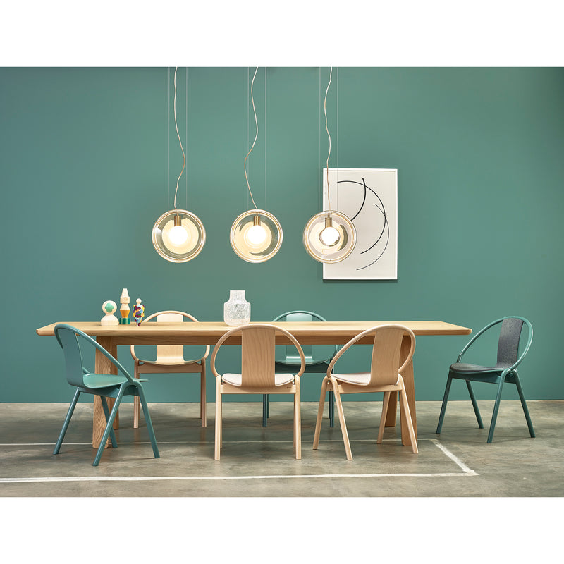 A modern dining room features a wooden table with mismatched stackable TON Chair Again Upholstered chairs against a teal wall. Three circular pendant lights hover above, minimalist artwork adorns the space, and concrete floors blend with neutral and teal tones for adaptable seating solutions.