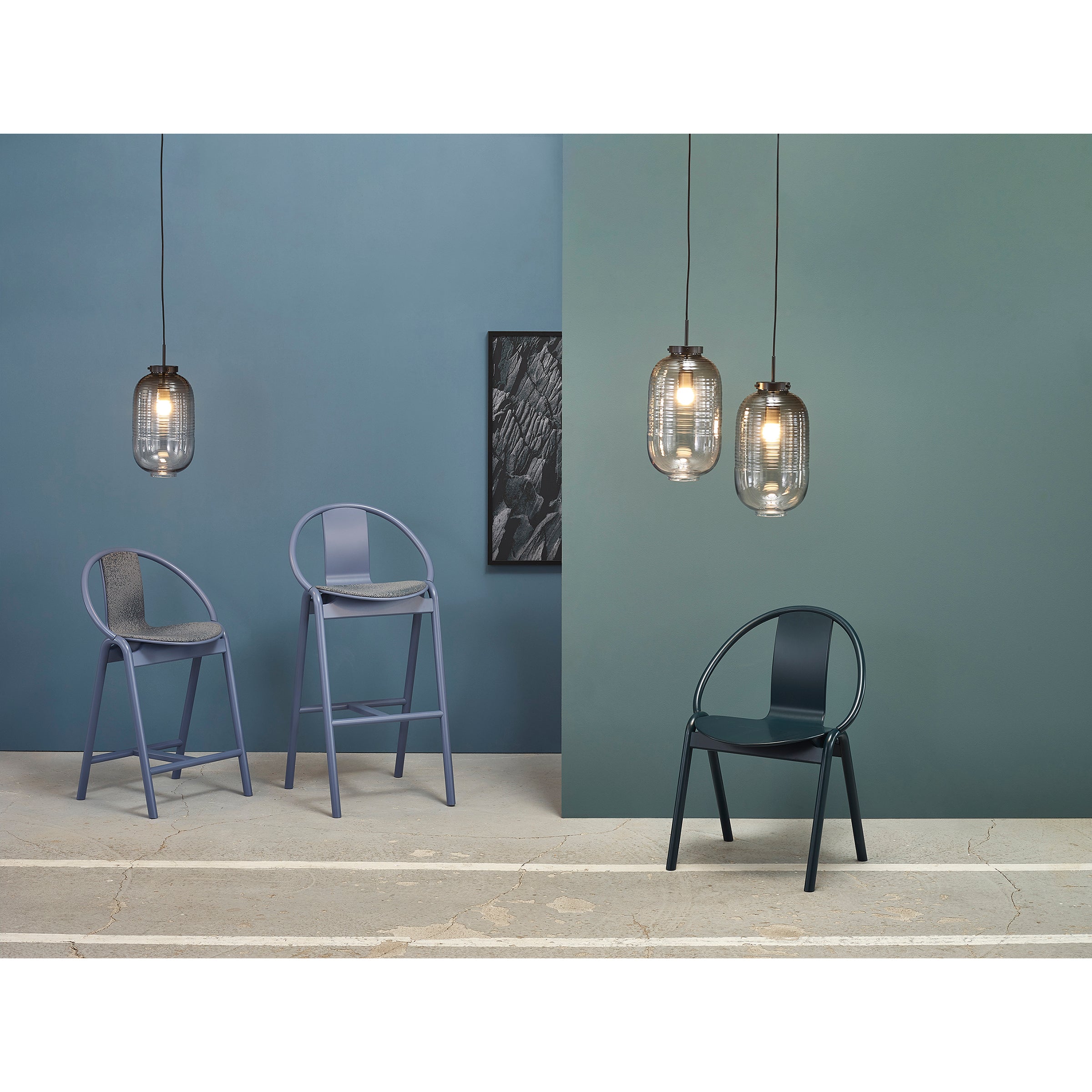 A minimalist room with a teal wall features three pendant lights and modern seating: two tall barstools, Chair Again Upholstered by TON in soft blue with gray cushions, made of solid beech wood, and one black Bentwood chair. A black-and-white framed artwork decorates the background.
