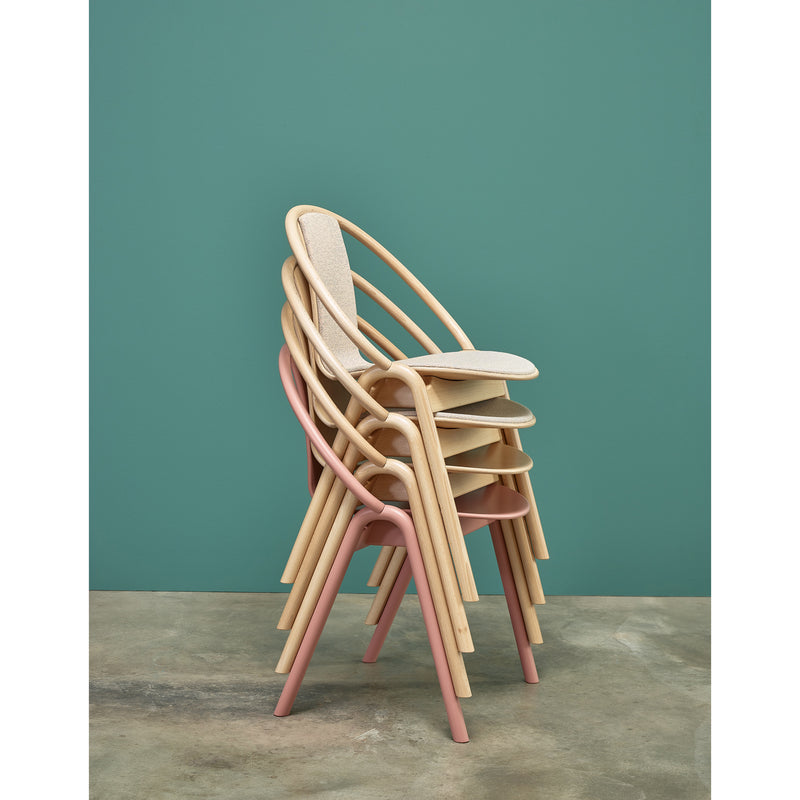 A stack of four Chair Again Upholstered dining chairs by TON is set against a green wall. Featuring wooden frames, the chairs have light fabric seats, pink front legs, and a beige frame. Neatly arranged on a concrete floor, they exemplify stylish design.