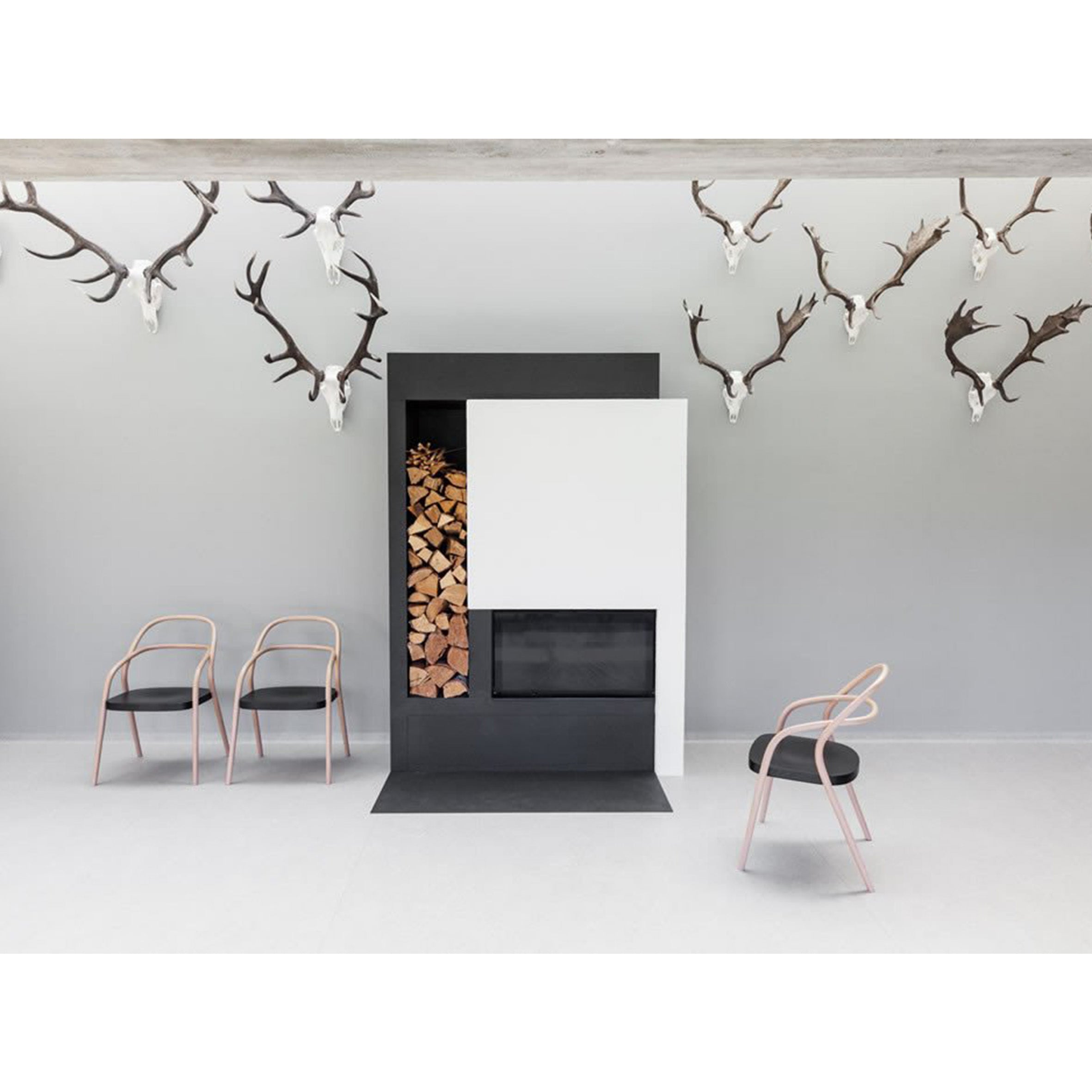 A modern minimalist room features a geometric black and white fireplace, stacked firewood, three light pink Chairs 002 - Upholstered from TON with black seats on the left, and multiple deer antlers mounted on the light gray wall.