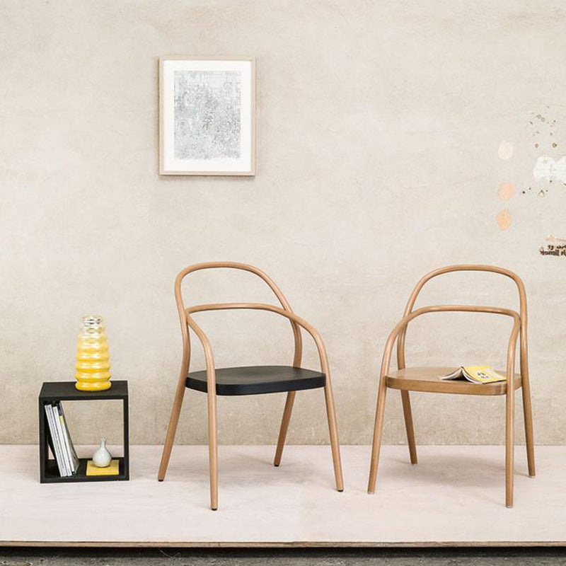 Two TON Chair 002s—minimalist wooden chairs, one with a black seat and another tan—are on a light platform. A black side table holds books, a vase, and a yellow object. Framed artwork decorates the textured beige wall. A small yellow booklet sits on the tan chair.