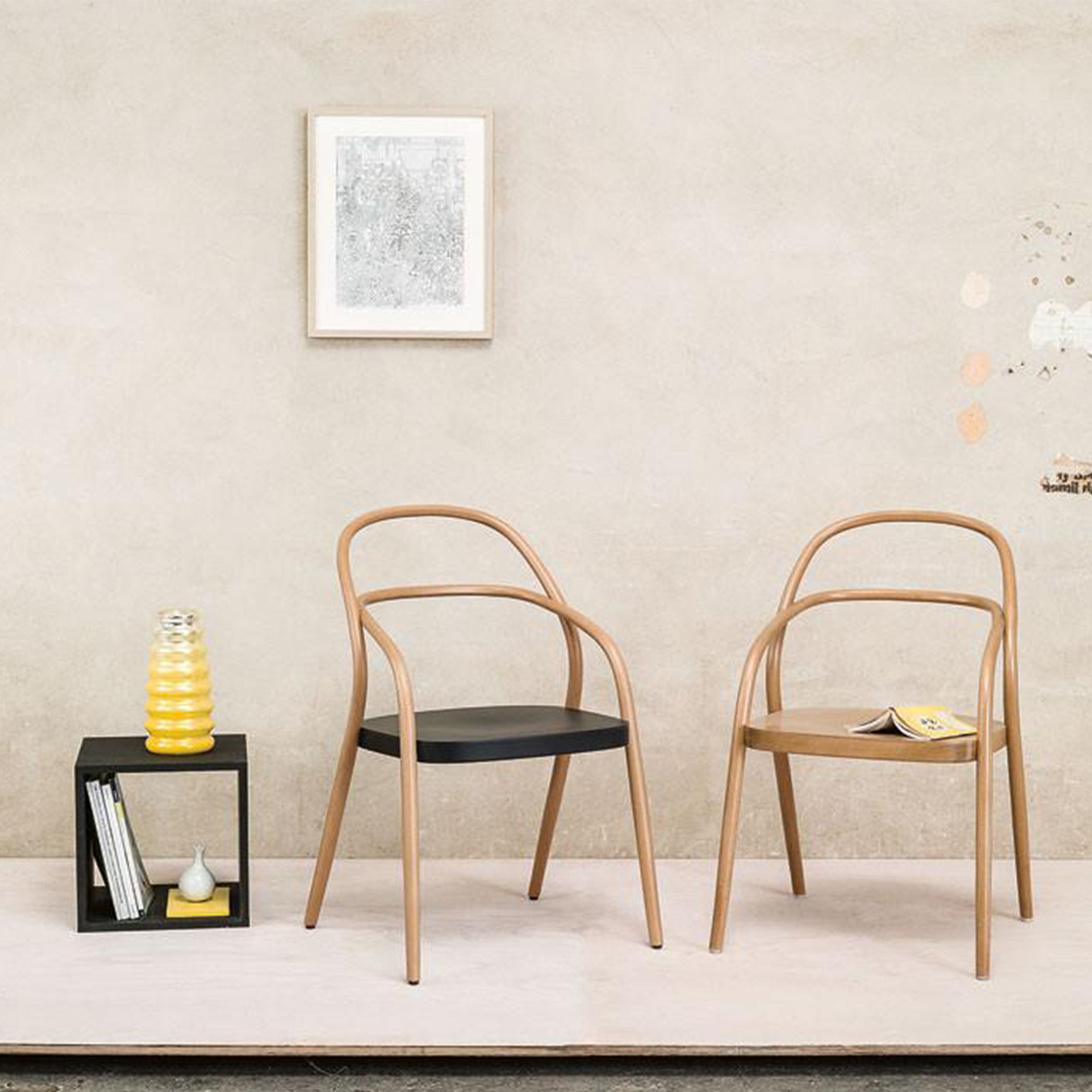 Two stylish Chair 002 - Upholstered pieces from TON stand before a beige wall. A small black side table holds a yellow vase and books, with a framed art piece above. A magazine rests on one chair, echoing the elegance of the No. 14 redesigns minimalist style and curved backrests by TONDesigner.