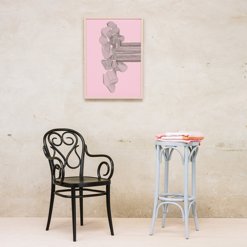 The black TON Armchair 04 with a curved design sits beside a light blue round table holding folded clothes. A framed pink abstract artwork hangs on the beige wall behind them.