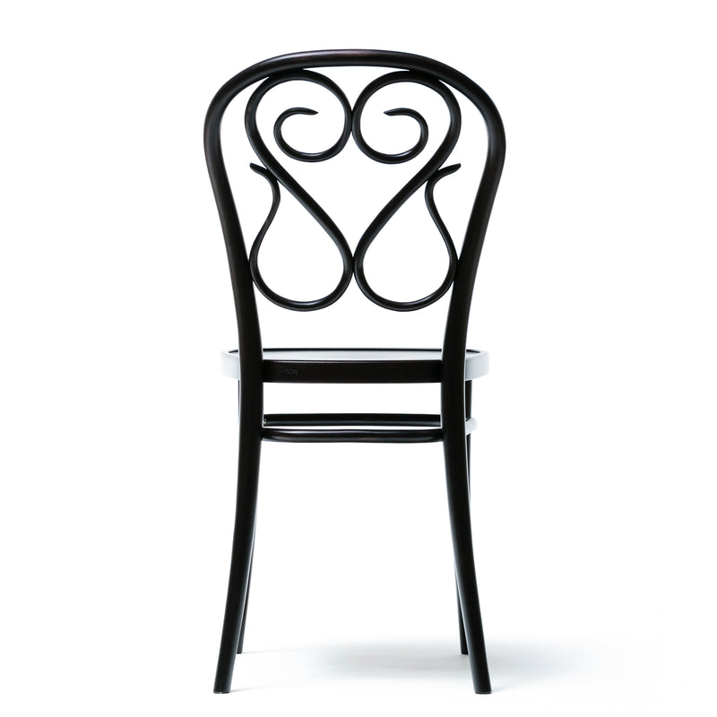 The Chair 04 by TON is a black, ornate dining chair with a decorative swirling backrest and slender legs. Its intricate bent details display elegant curves and loops, while the plain white floor and background accentuate its artistic features as seen from the rear.