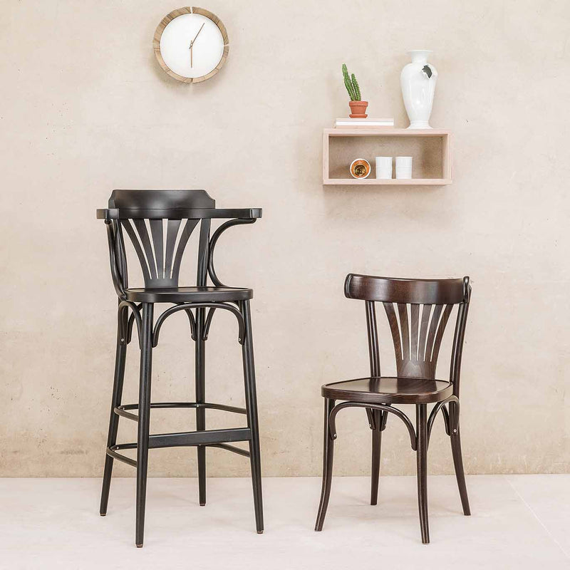 Two black wooden chairs, including a TON Barstool 135 with armrests, are set against a beige wall. Above them is a round clock and pink shelf with a white vase, cactus, and cup. The minimalistic setting features modern plywood design elements.