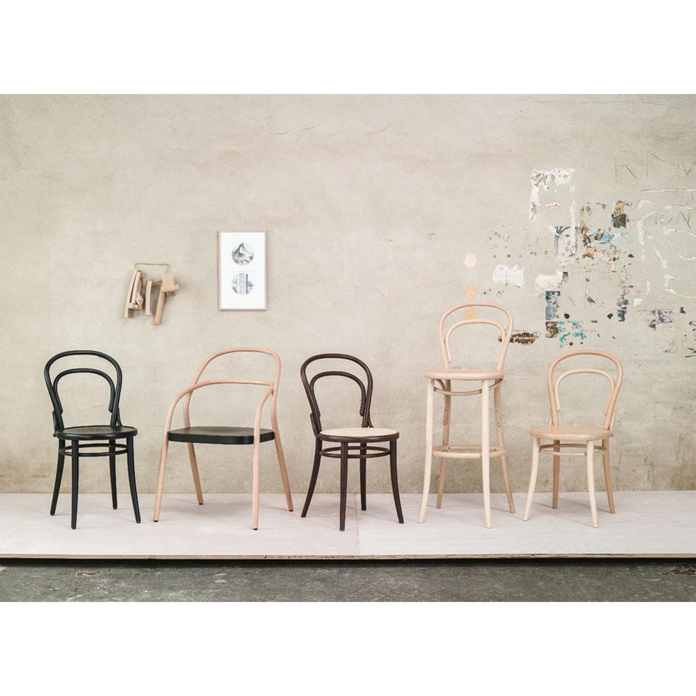 A row of five wooden chairs in different styles and colors is displayed against a textured wall. Among them is Chair 14 - Upholstered by TON, featuring beech rods. The backdrop includes a picture frame and paint marks, while the floor is made of light wood.