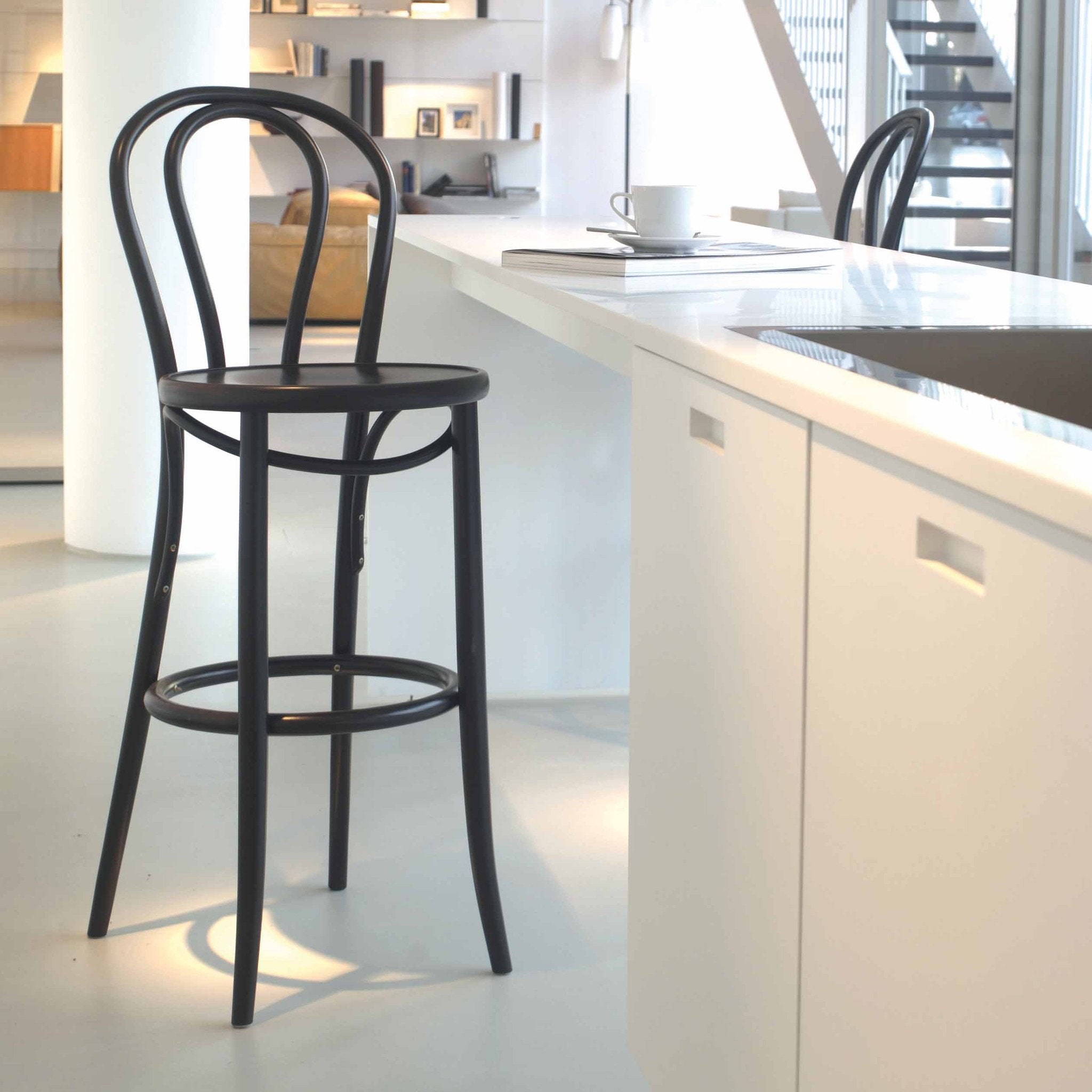 A modern kitchen features a sleek white counter and TONs Barstool 18, with an upholstered curved backrest. Soft lighting highlights the minimalist design, while plywood shelves and stairs create a warm backdrop.