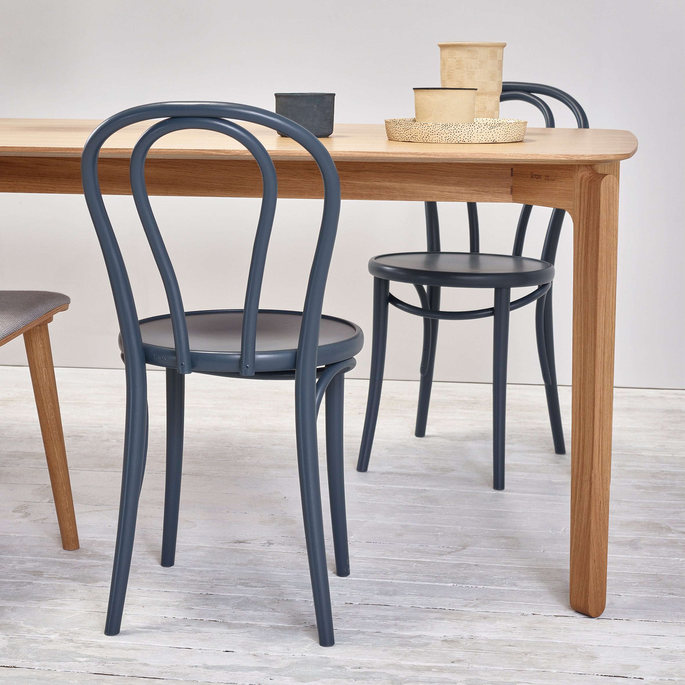 A light wood table with a beech-like finish sits on a light wooden floor. Around it are two black Chair 18 - Upholstered by TON. On the table are two ceramic cups, a woven basket, and a small vase.
