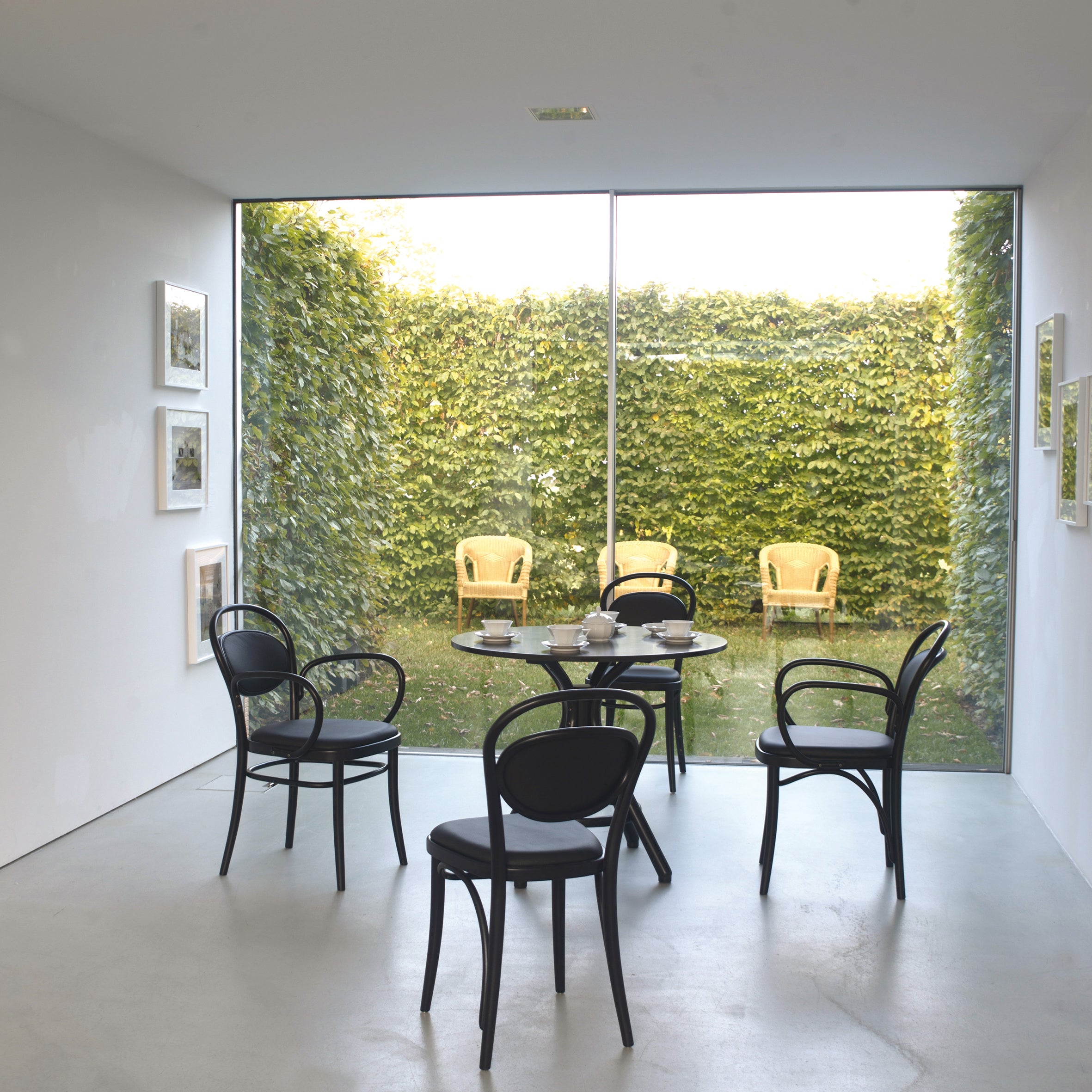 A minimalist dining area features a glass table with black chairs showcasing beech finishes on a light floor. By the large window, a TON Armchair 20 complements the view of a lush courtyard with four light-colored chairs. Framed art adorns the walls, and natural sunlight fills the space.
