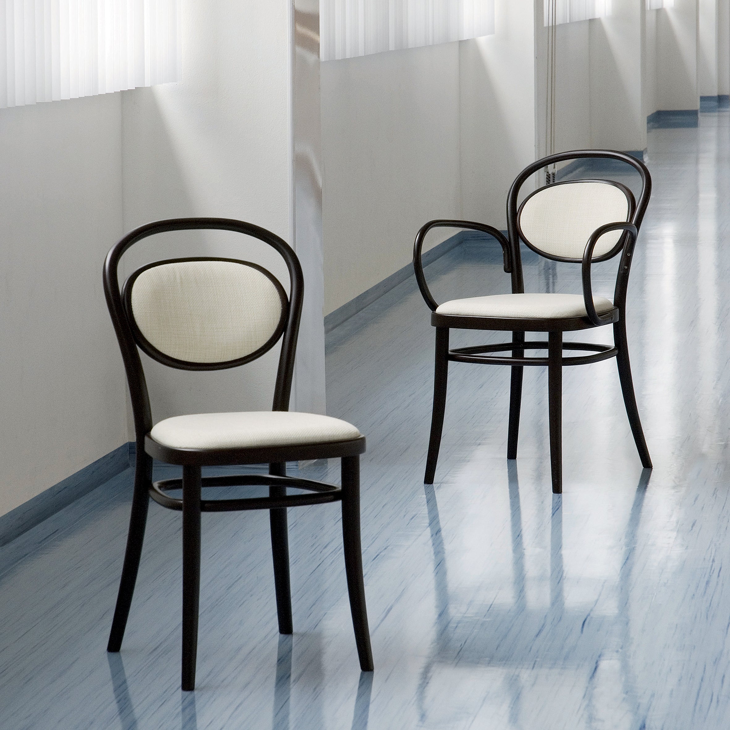Two elegant TON Chair 20 - Upholstered dining chairs, featuring dark wooden frames and light upholstered seats, are situated in a well-lit hallway with light blue flooring. The modern and minimalist space is softly illuminated by natural lighting through sheer curtains.