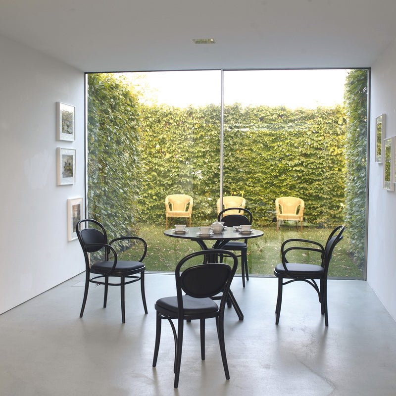 A modern dining room features a round glass table and four black dining chairs. A large window showcases a green outdoor area with three wicker chairs with cane seating, while minimalist white walls are adorned with small framed artworks. The black chairs could be similar to Chair 20 - Upholstered by TON.