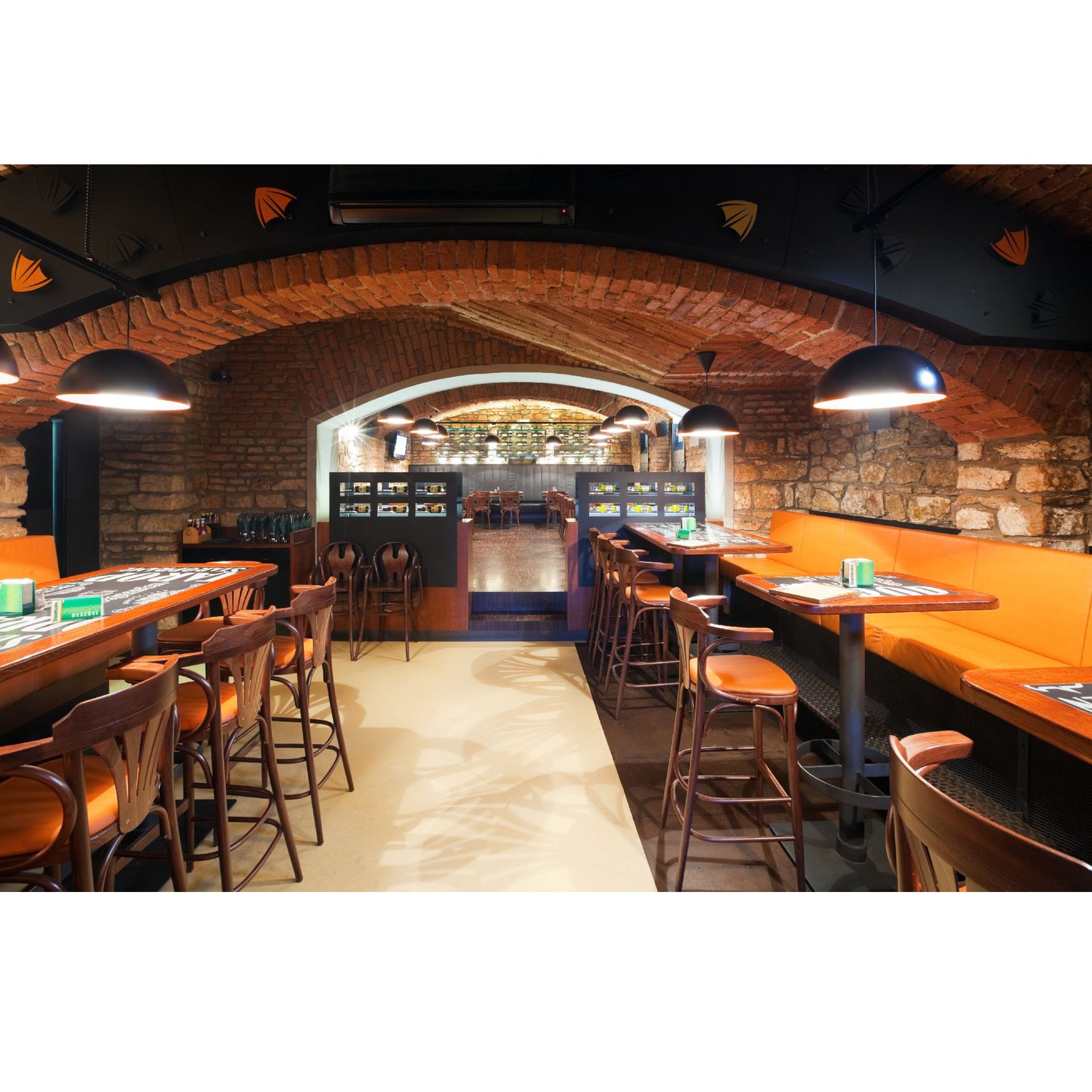The cozy restaurant features an arched brick ceiling and stone walls. Wooden tables, chairs, TONs Armchair 25, and orange cushioned bench seating line the narrow walkway. Overhead lamps provide soft lighting, creating a warm ambiance.