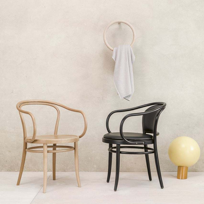 Two elegant TON Armchair 30s, one beige and one black, grace a light wooden floor by a neutral wall. Their open backrest design adds charm. A white towel hangs from a circular peg above, while a round yellow lamp sits nearby on the floor, showcasing their versatility.