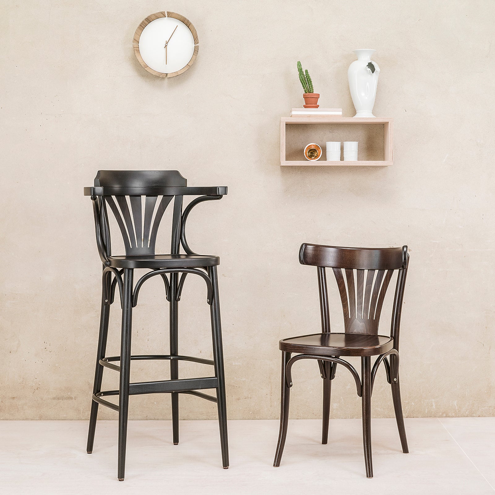 A minimalist room features a TON Chair 56 with a plywood fan backrest and curved design, along with a tall black bar stool. A wall clock hangs above, while a small shelf holds a white vase and cactus against a light beige wall for an elegantly simple look.