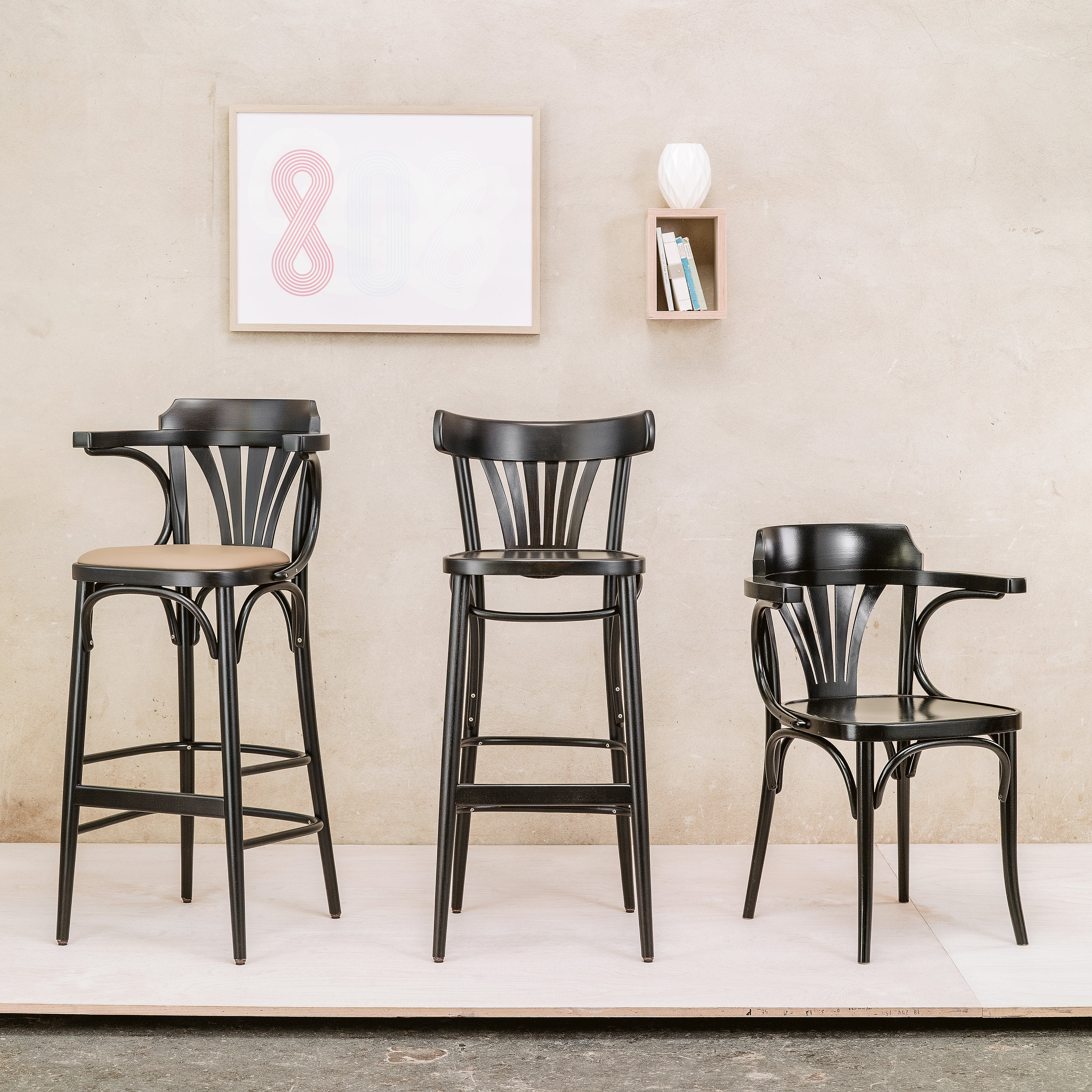 Three black wooden chairs, including the upholstered dining Chair 56 by TON, are arranged on a white platform against a beige wall. Above them, a framed abstract art piece and a small shelf with books and a white vase add flair to the scene.