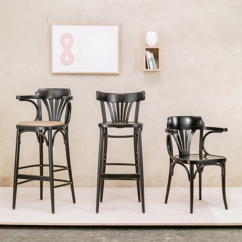 Three black wooden chairs, including the upholstered dining Chair 56 by TON, are arranged on a white platform against a beige wall. Above them, a framed abstract art piece and a small shelf with books and a white vase add flair to the scene.