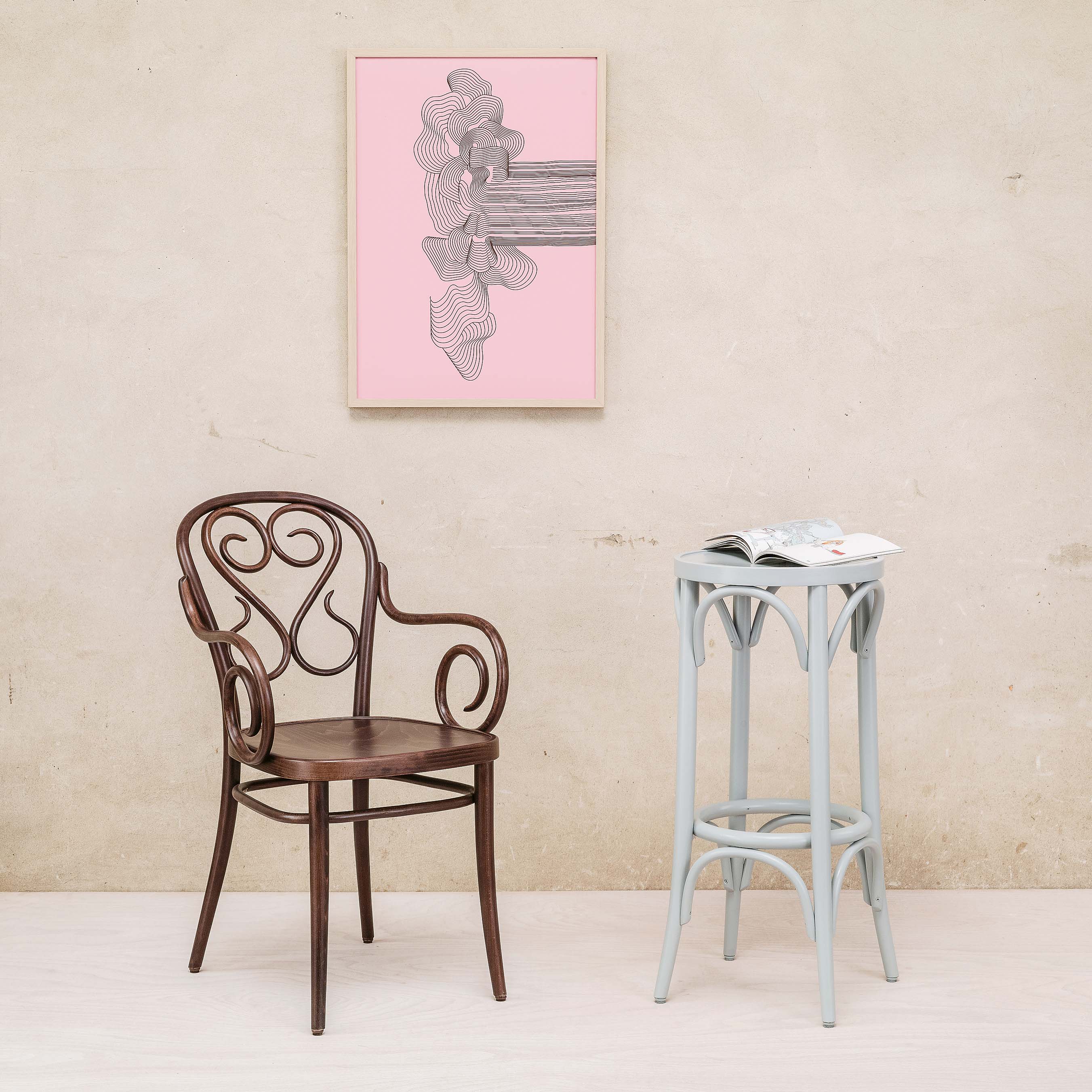 A dark wood chair with a swirling backrest stands next to the TON Barstool 73, which is light blue and accompanied by an open magazine. A framed black and white abstract artwork on a pink background decorates the beige wall, bringing charm to the beech-tone room.