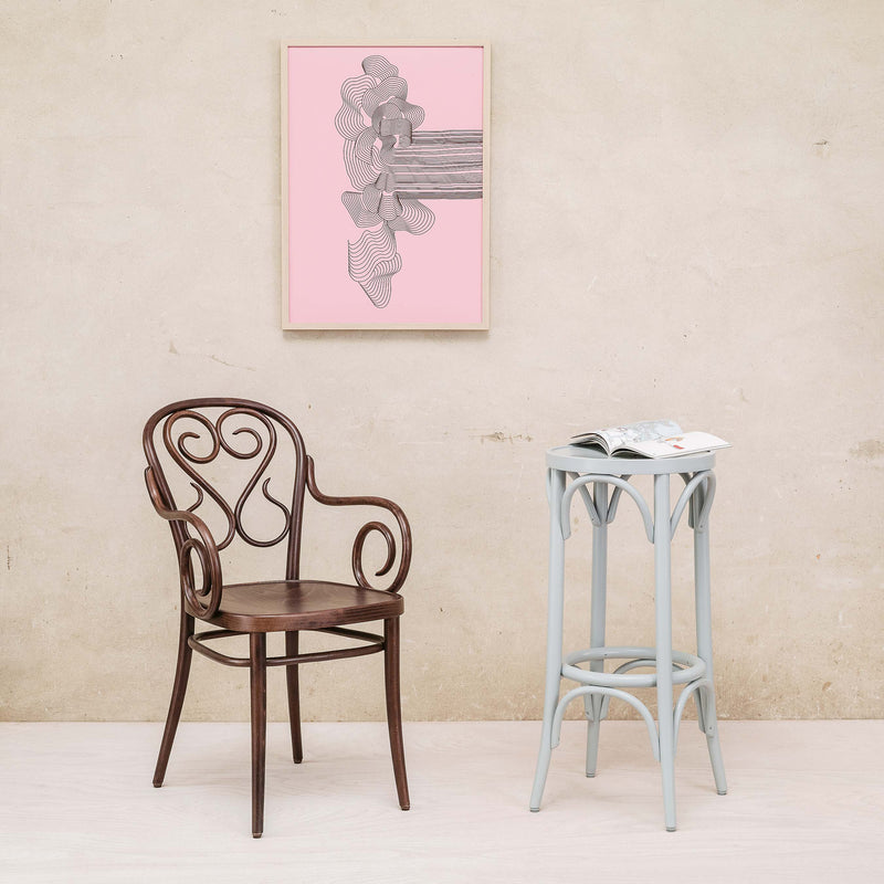 A dark wood chair with a swirling backrest stands next to the TON Barstool 73, which is light blue and accompanied by an open magazine. A framed black and white abstract artwork on a pink background decorates the beige wall, bringing charm to the beech-tone room.