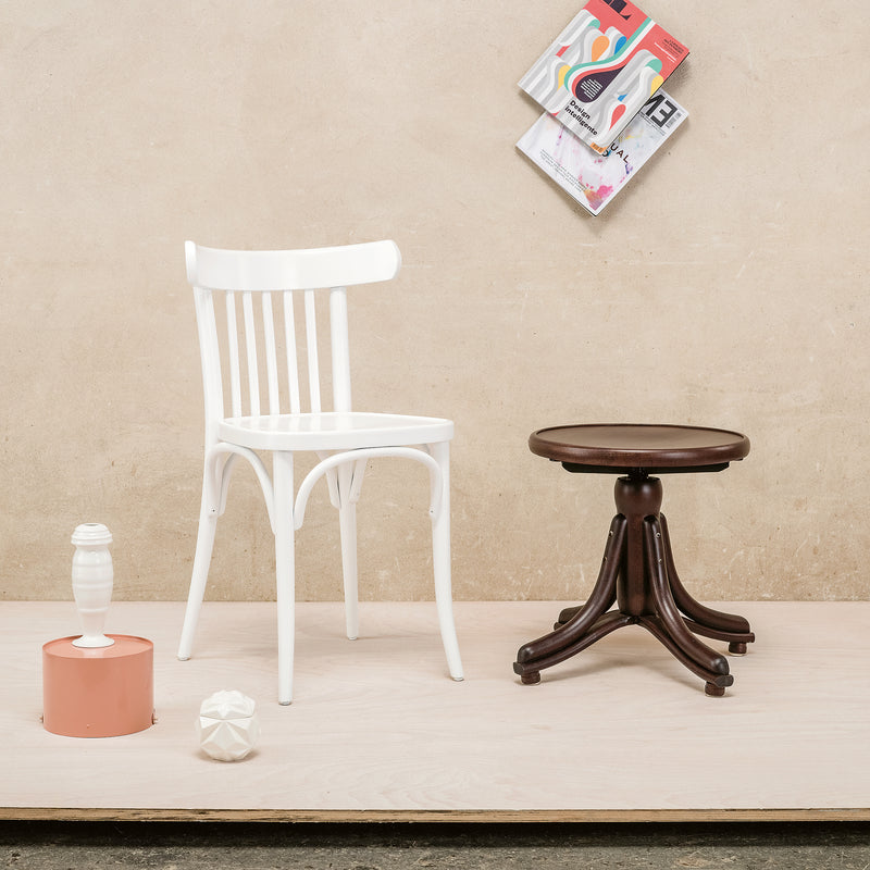 A white Chair 763 by TON, crafted from beech sits on a light wooden floor alongside a brown round stool. A pink cylinder, a white vase, and a geometric ornament add decor. Above, two magazines are arranged in a staggered modern interior style.