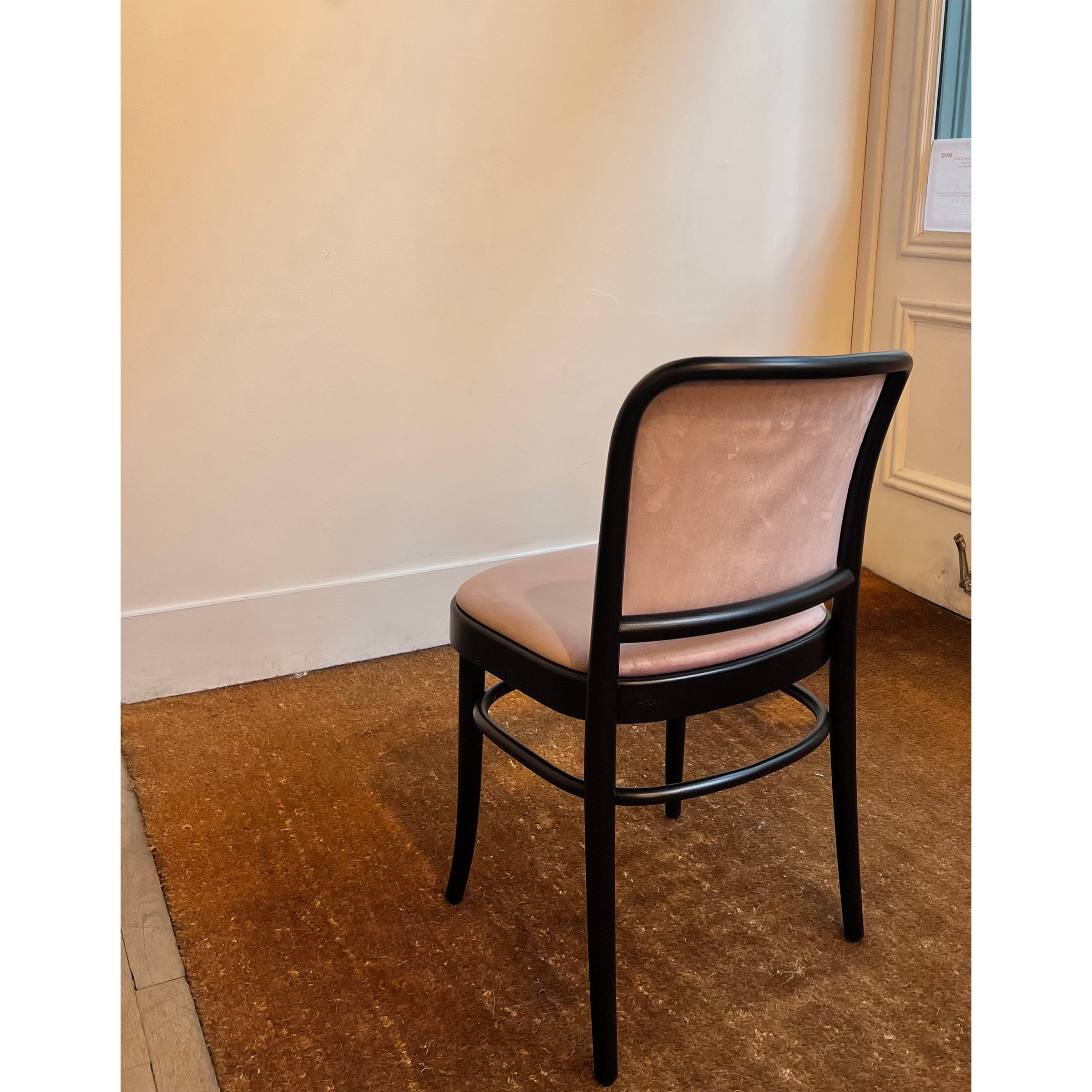 A single TON Chair 811 with a dark frame and light upholstery reminiscent of Josef Hoffmann design sits on a brown carpet against a pale wall with a high baseboard, near a door adorned with white paper. The setting is simple and serene, evoking Art Nouveau elegance.