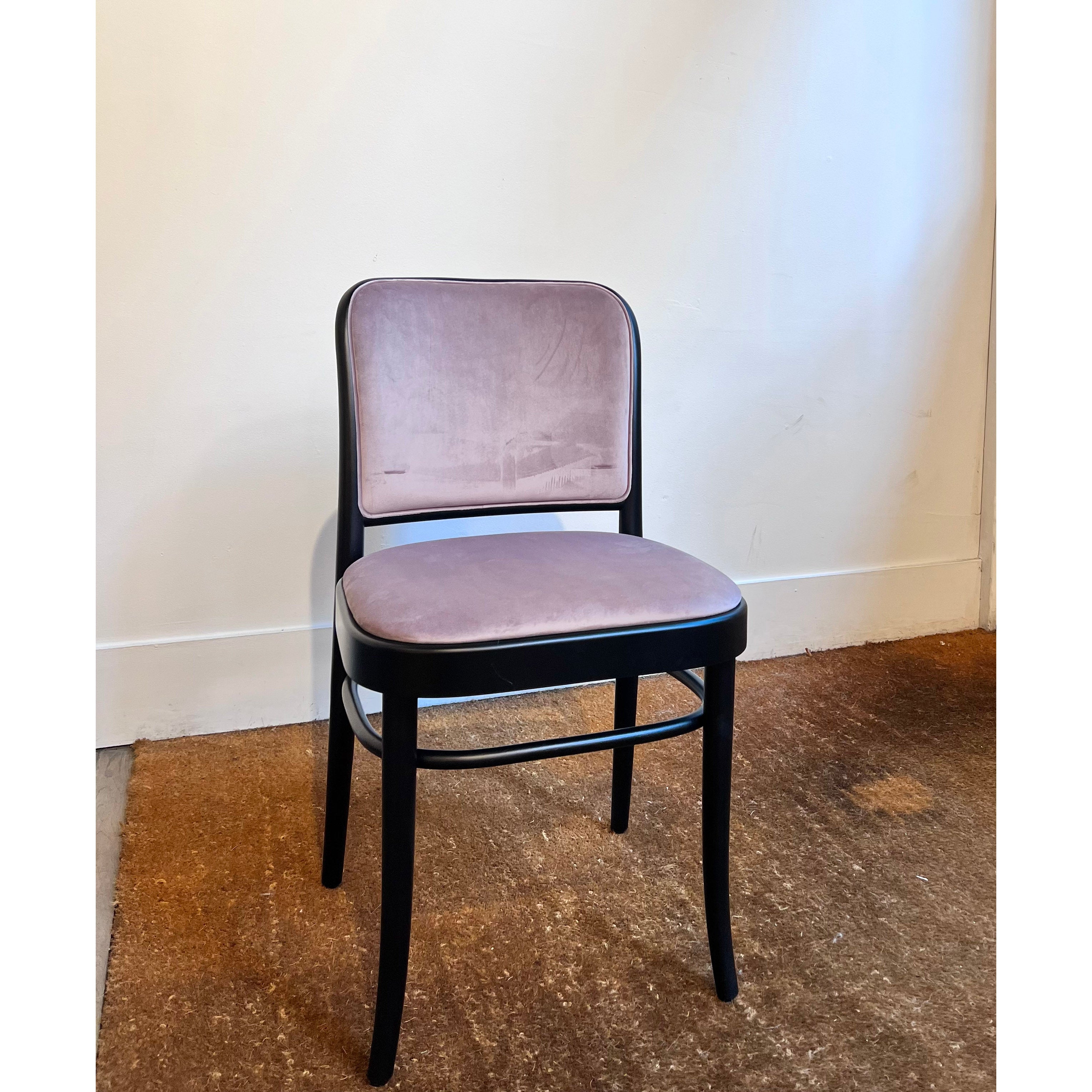 A Chair 811 by TON, featuring a simple design, rests against a white wall. Its light purple cushioned seat and backrest evoke Josef Hoffmanns Art Nouveau style. The brown floor beneath shows slight wear.