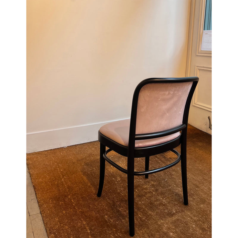 A single TON Chair 811 with a dark frame and light upholstery reminiscent of Josef Hoffmann design sits on a brown carpet against a pale wall with a high baseboard, near a door adorned with white paper. The setting is simple and serene, evoking Art Nouveau elegance.