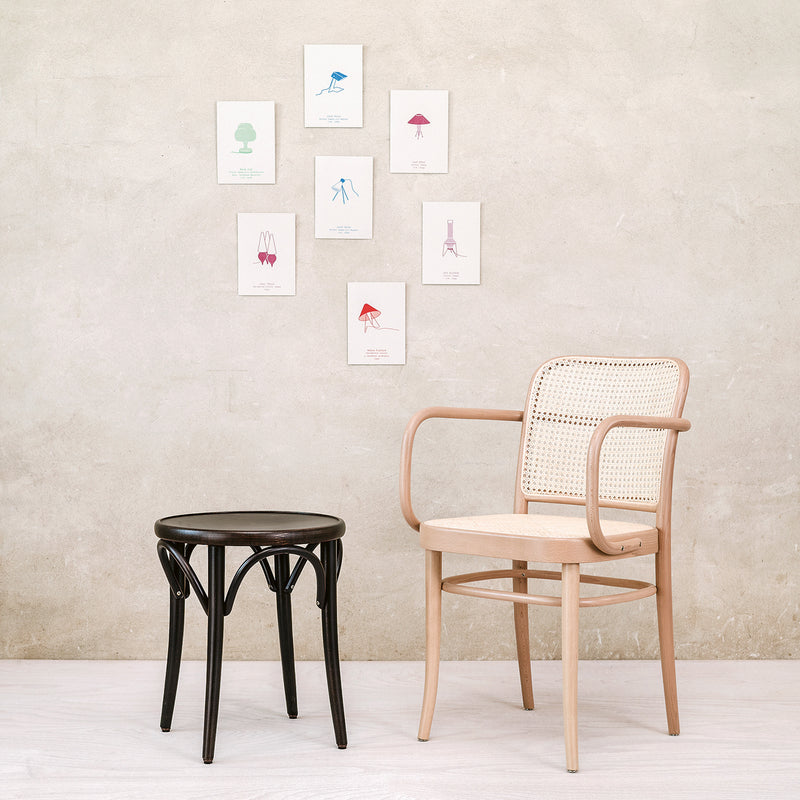 A minimalist room with a beige wall showcases six small framed prints in a grid. In front is TONs Armchair 811 with a woven back inspired by Josef Hoffmann, next to a dark round wooden stool. The light-colored floor enhances the serene Art Nouveau ambiance.
