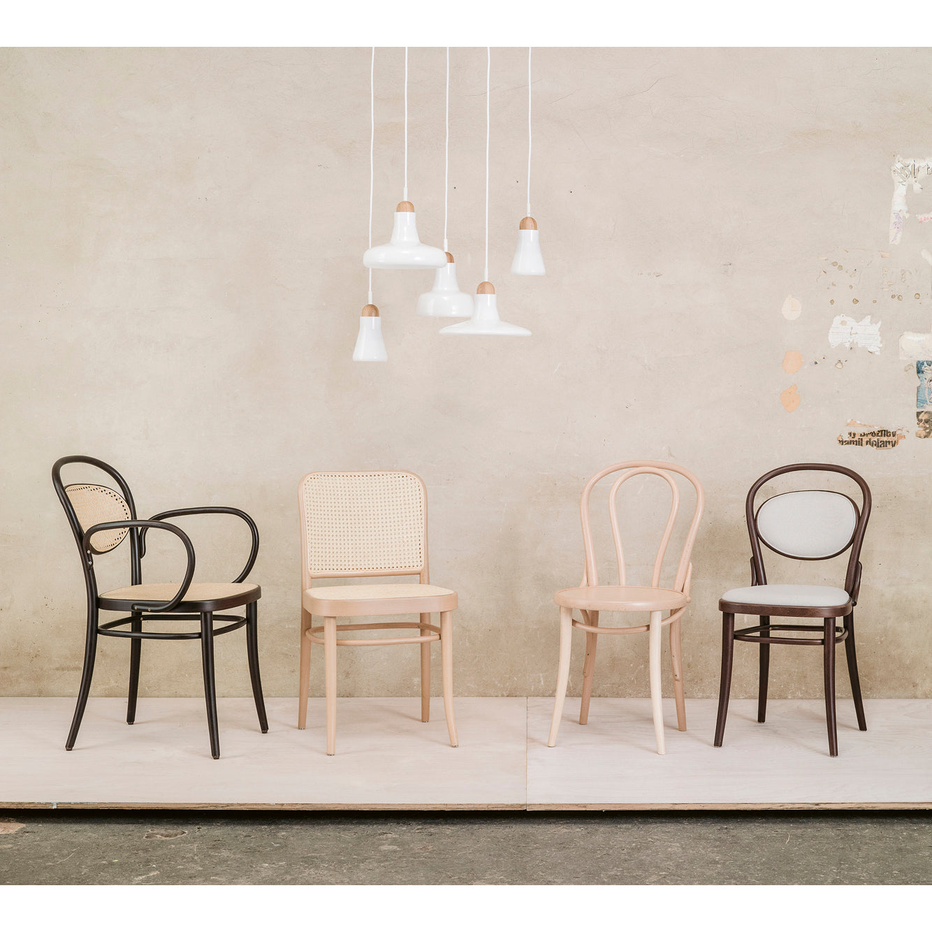Four TON Chair 811 - Cane wooden dining chairs, inspired by Josef Hoffmann design, are lined up against a beige wall. Three white pendant lights above create a soft glow. The scene is minimalistic with a neutral palette, reflecting Art Nouveau elegance.